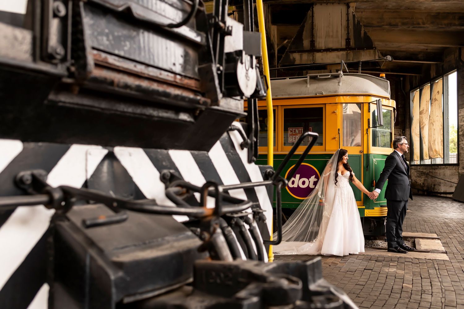 Couple poses near vintage train in industrial railway setting.
