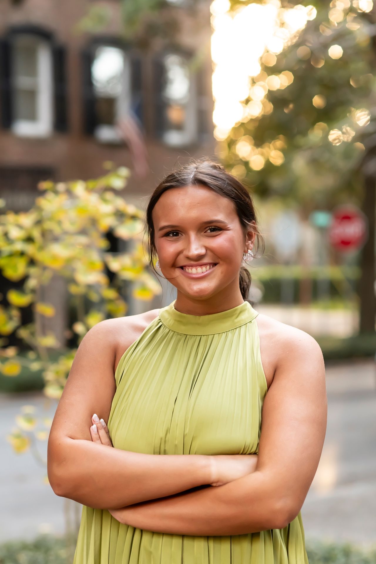 A person in a lime green halter dress stands with arms crossed, smiling outdoors with blurred bokeh lighting in background.