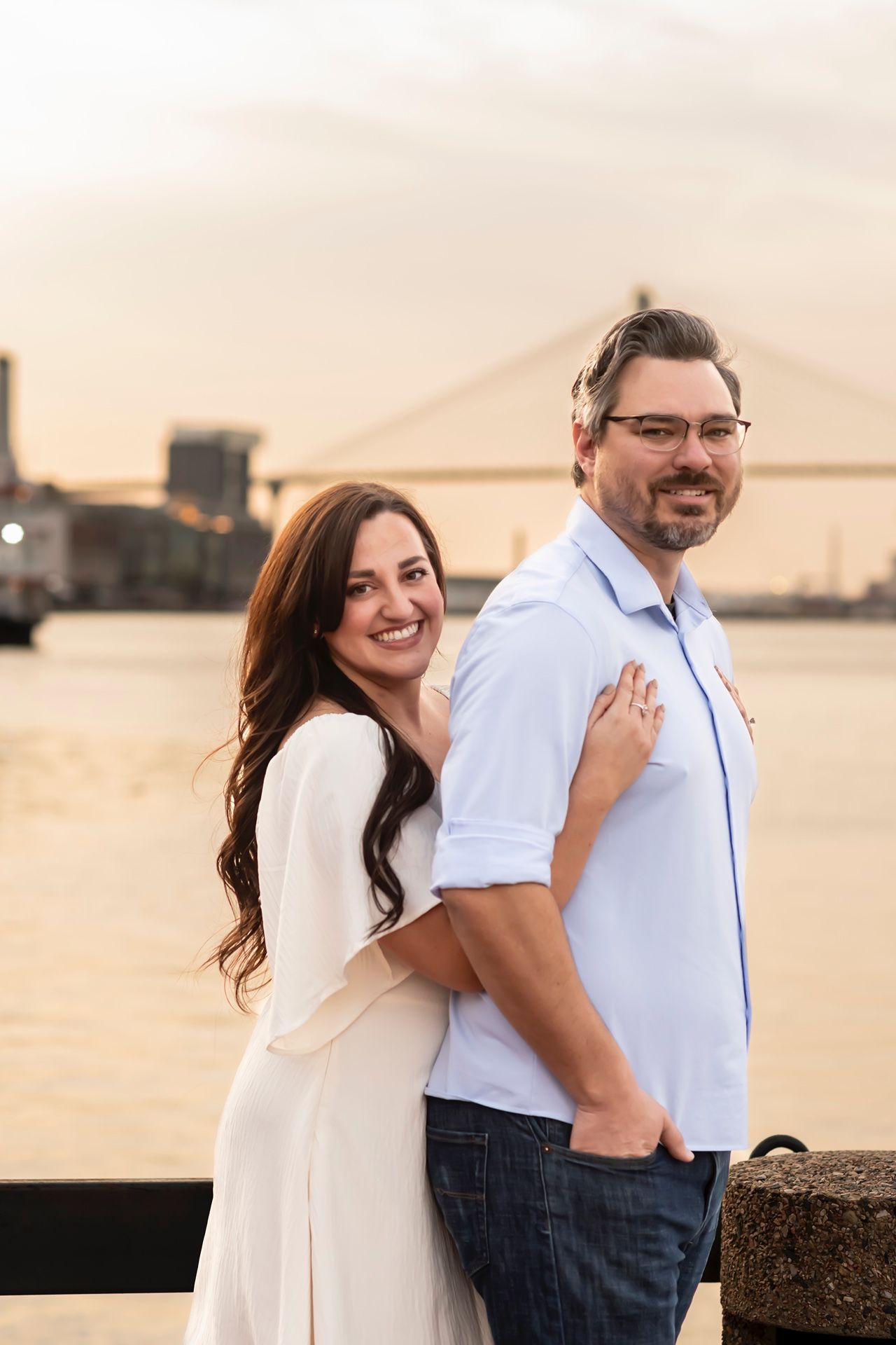 Couple stands together on waterfront at sunset with bridge silhouette in background.