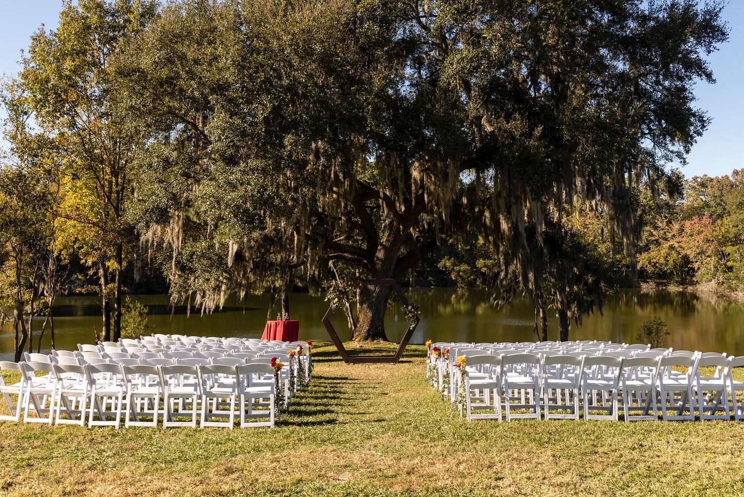 White wedding chairs set up on a lawn beside a lake with Spanish moss draped oak trees and red archway decoration.