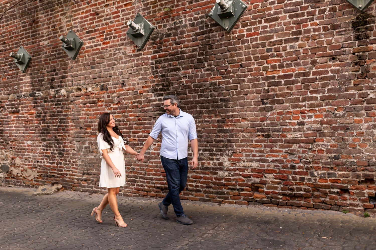 Couple walks together along historic red brick wall with security lights mounted above in urban setting.