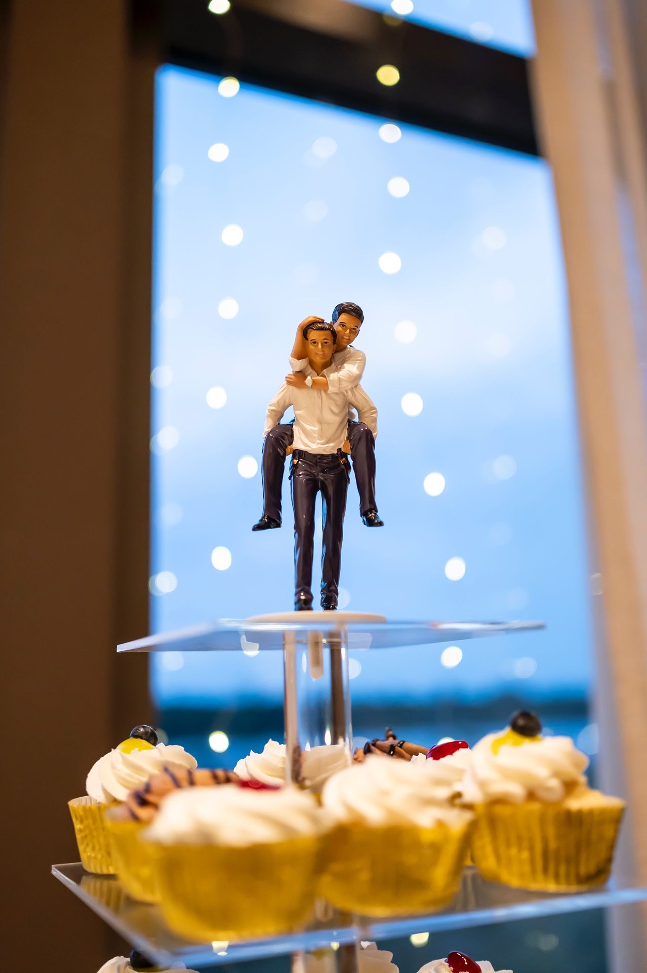 A wedding cake topper displays a romantic couple on a glass dessert stand with cupcakes arranged below against a bokeh background.