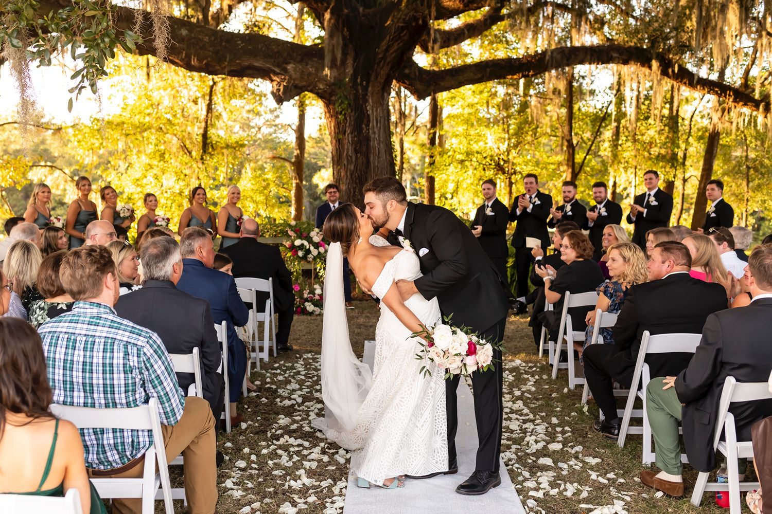 A couple shares their first kiss during an outdoor wedding ceremony under a large oak tree draped with Spanish moss.