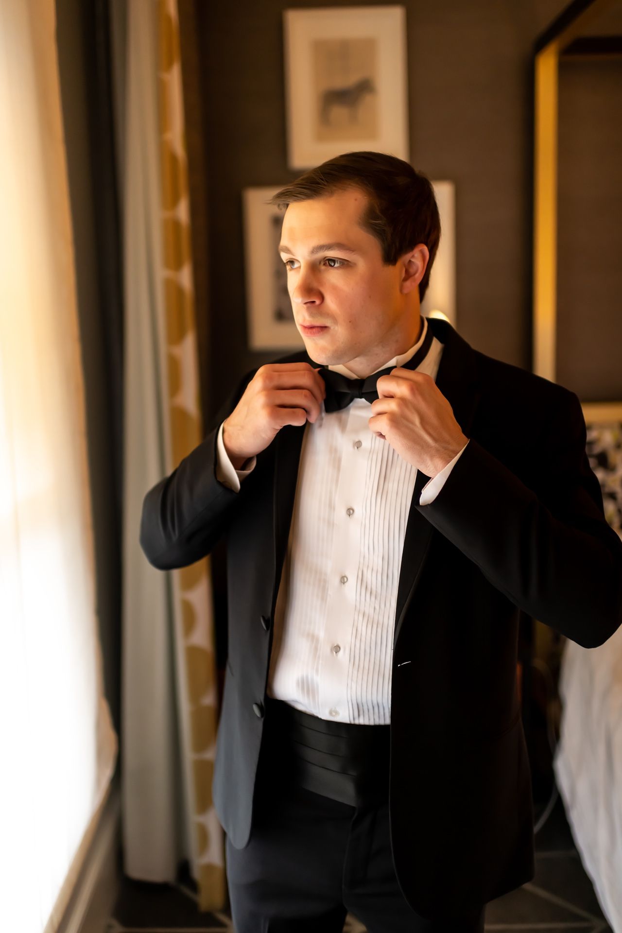 A person in a formal tuxedo and white dress shirt adjusts their bow tie while looking in mirror.
