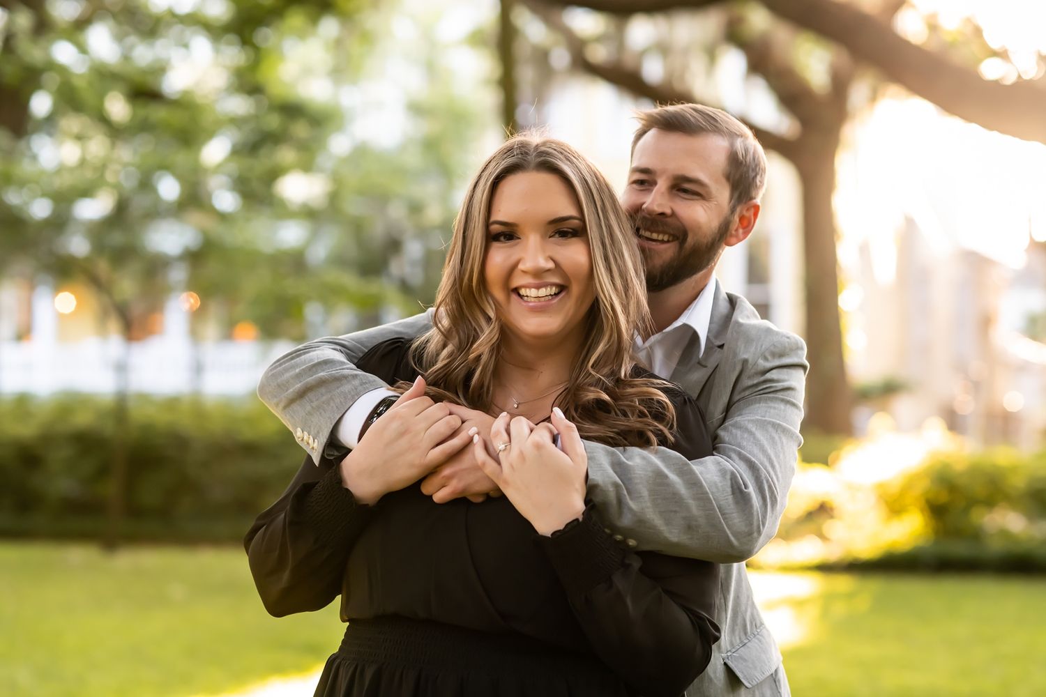 A couple embraces and laughs together in a sunny park with green trees in the background.