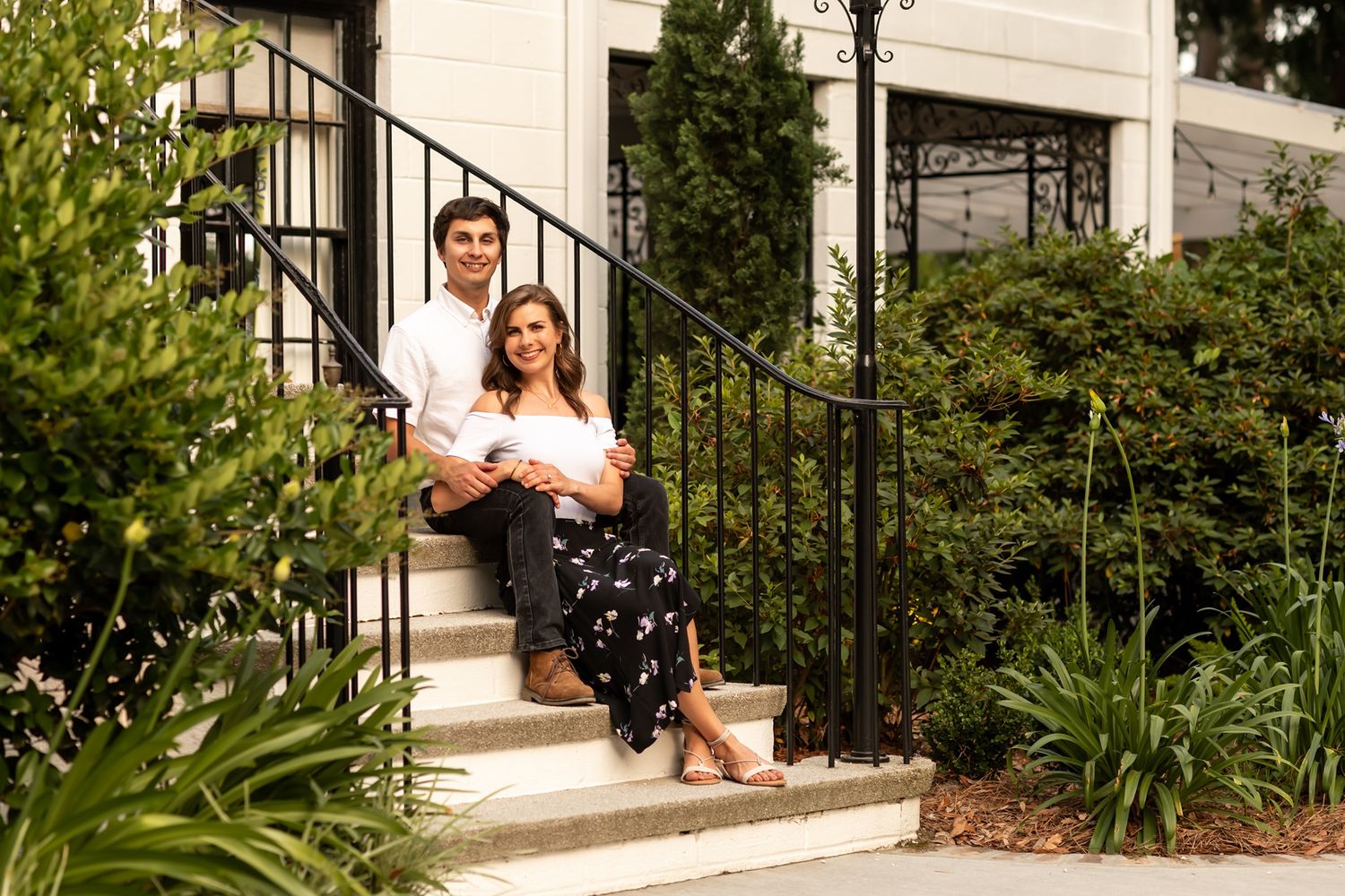 Two people sitting together on white concrete steps with black metal railings and green landscaping around them.