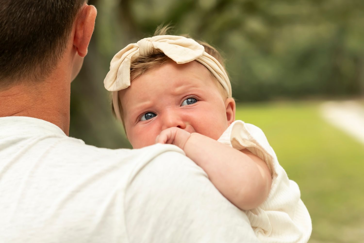Baby in cream-colored outfit rests on shoulder while looking back at camera with bright eyes and bow head accessory.