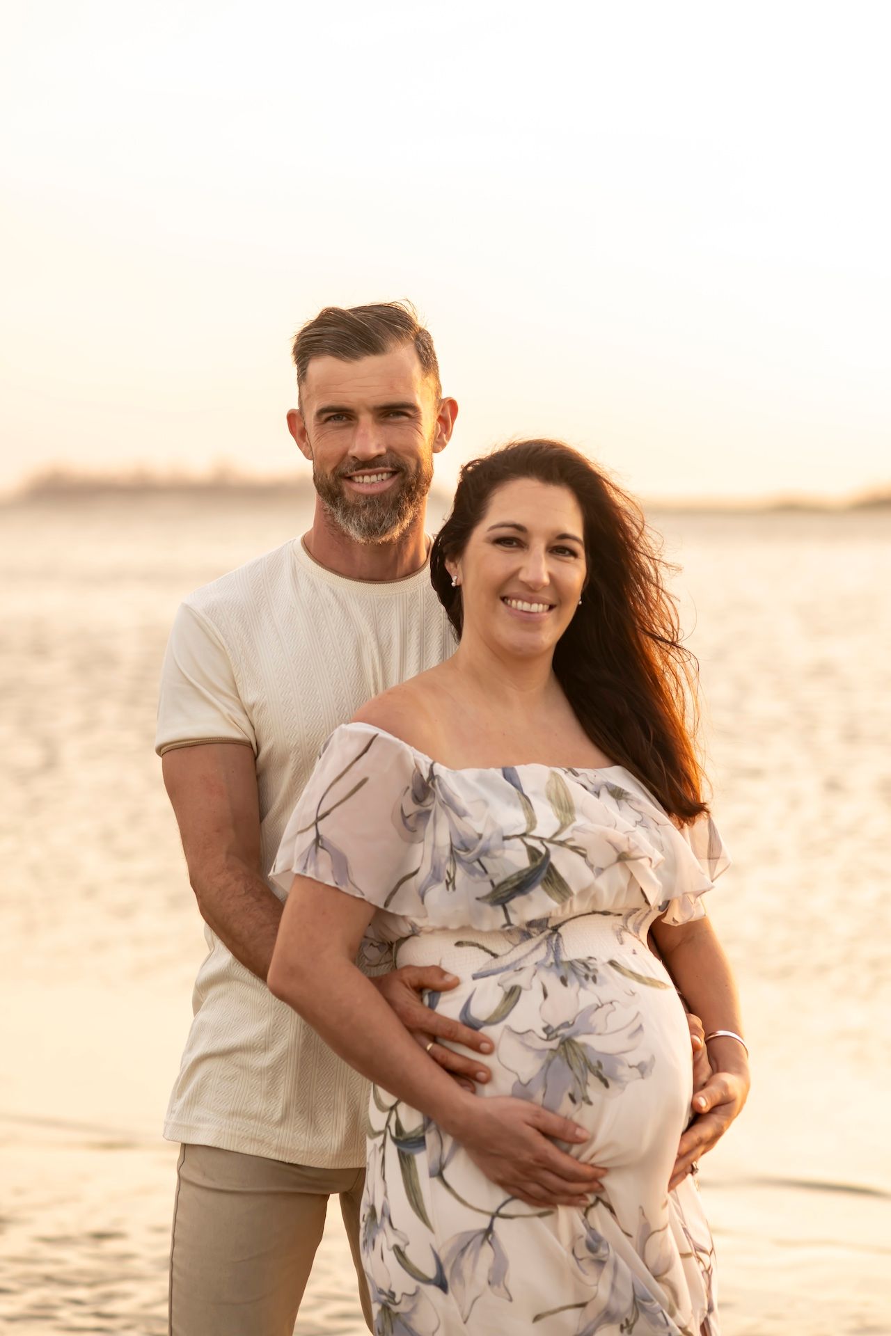 A couple stands together on a beach at sunset while cradling a pregnant belly in a floral dress.