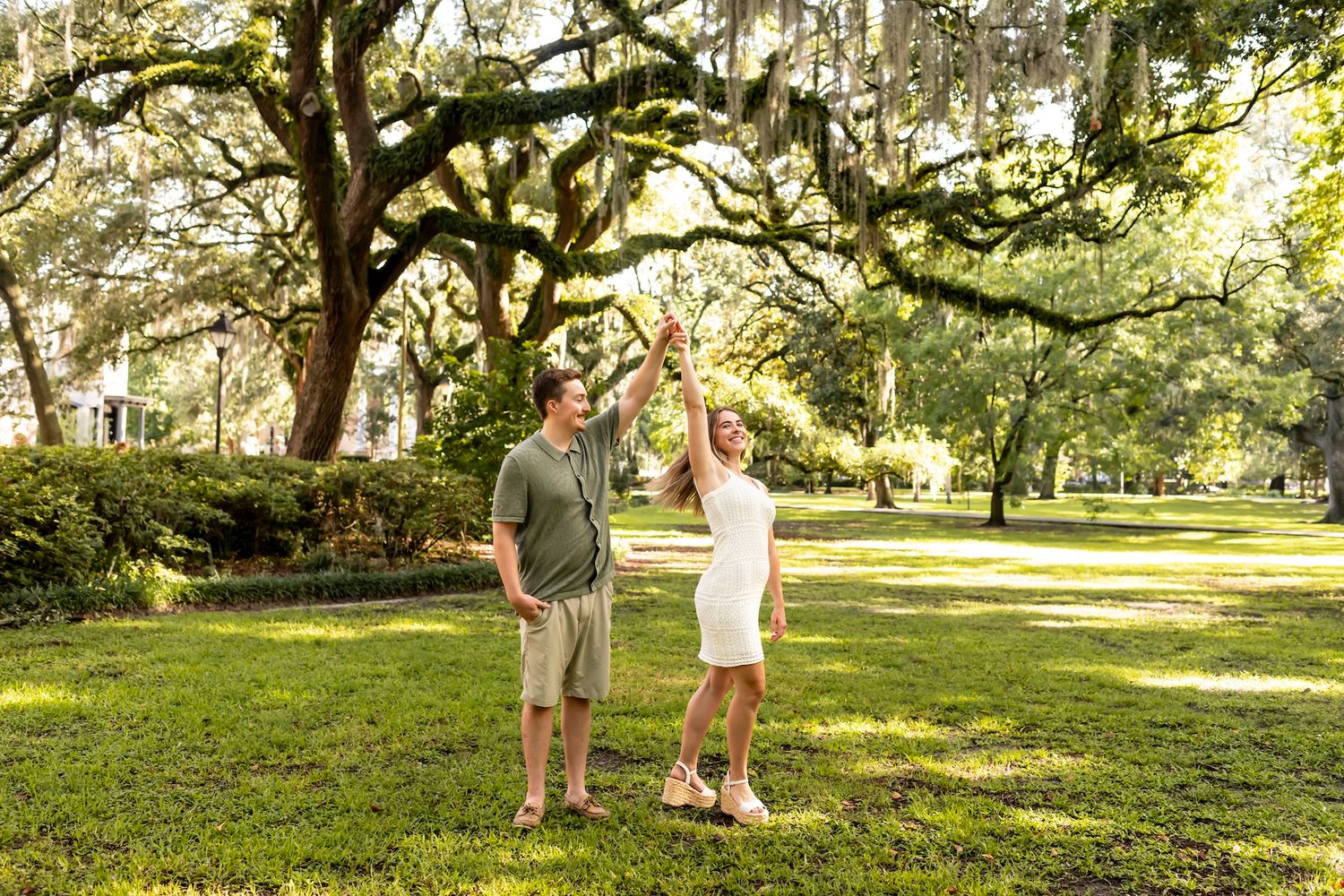 Two people stand together under moss-draped oak trees in a scenic park garden on a sunny day.