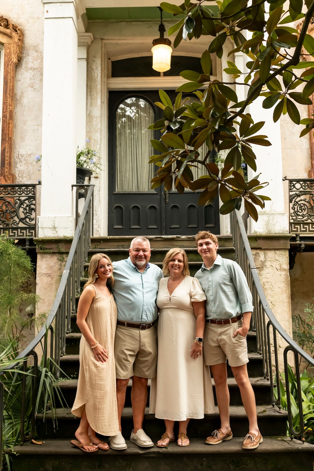 Group stands on steps of historic southern home with vintage door, lush magnolia leaves, and decorative railings.