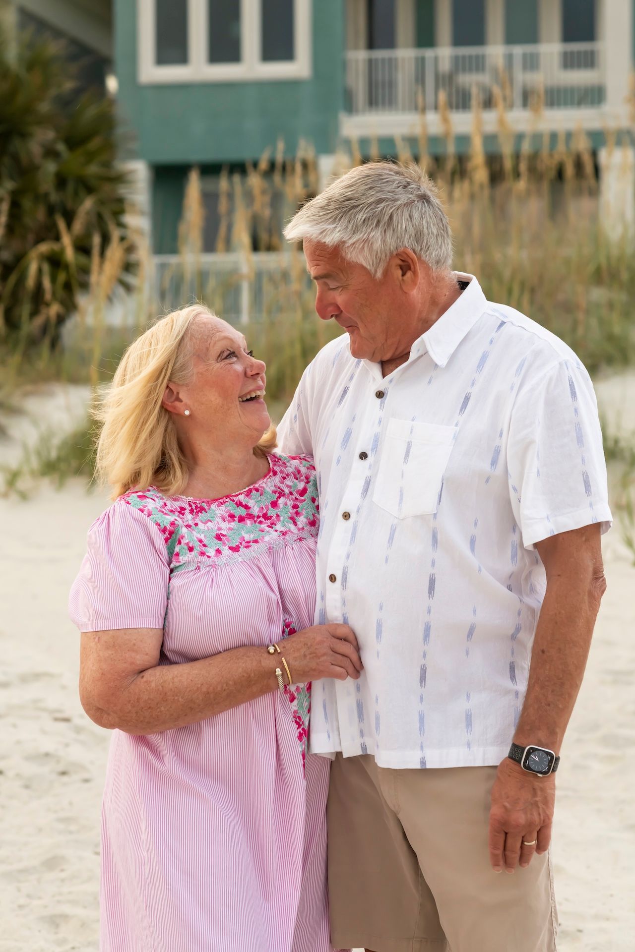 Couple sharing joyful laughter together on a sunny beach while wearing coordinated pink and white summer outfits.