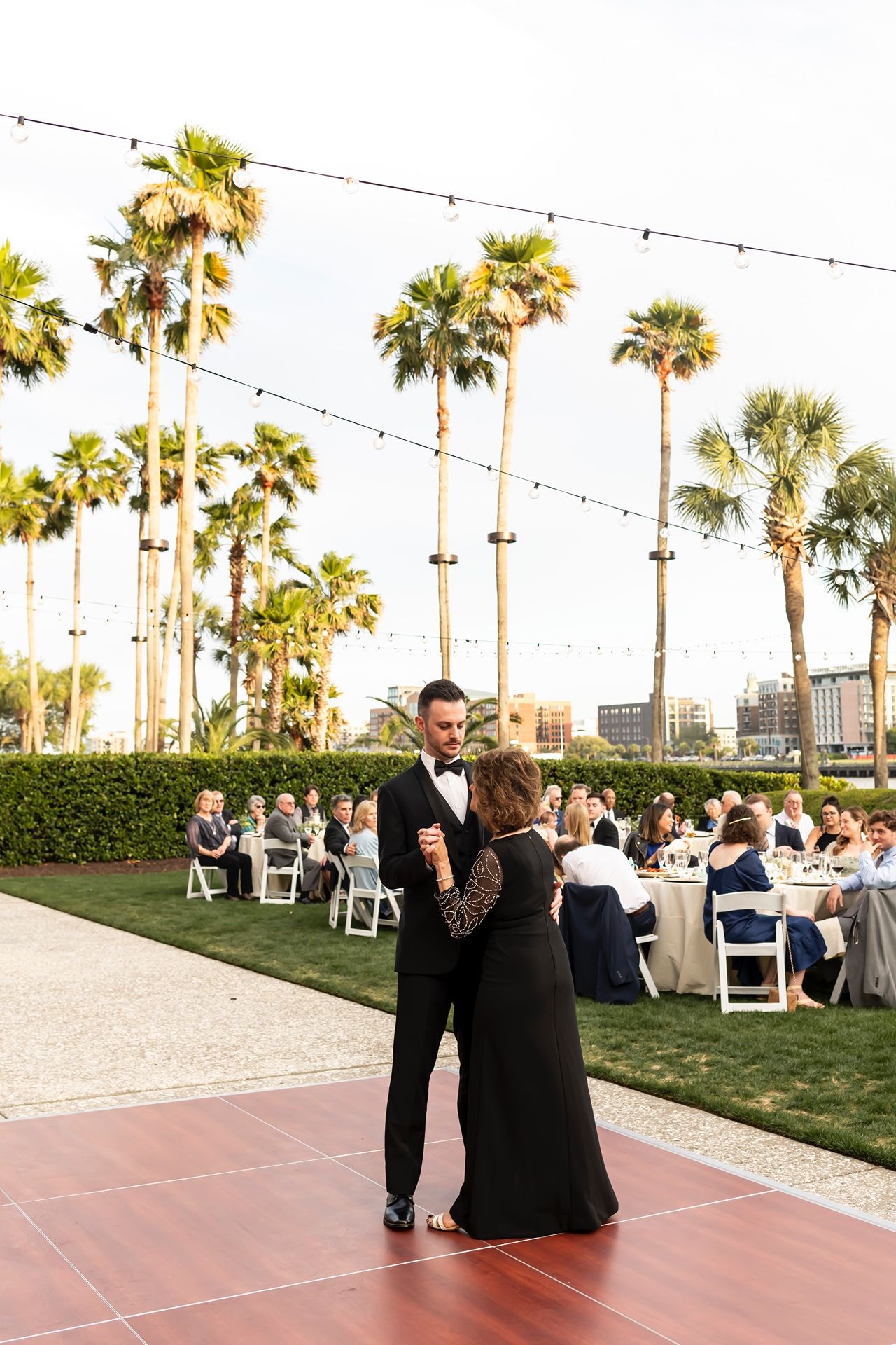A wedding reception at an outdoor venue with palm trees and string lights while a couple dances on the red tile floor.