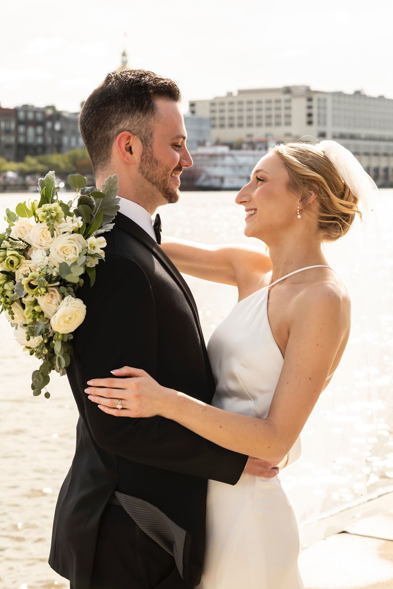 A happy couple in formal wedding attire dance and embrace outdoors along a waterfront with urban buildings in background.