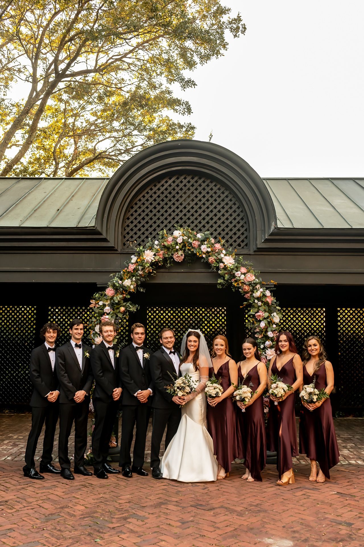 A wedding party in formal attire poses elegantly against a dark lattice backdrop with floral arch decorations.