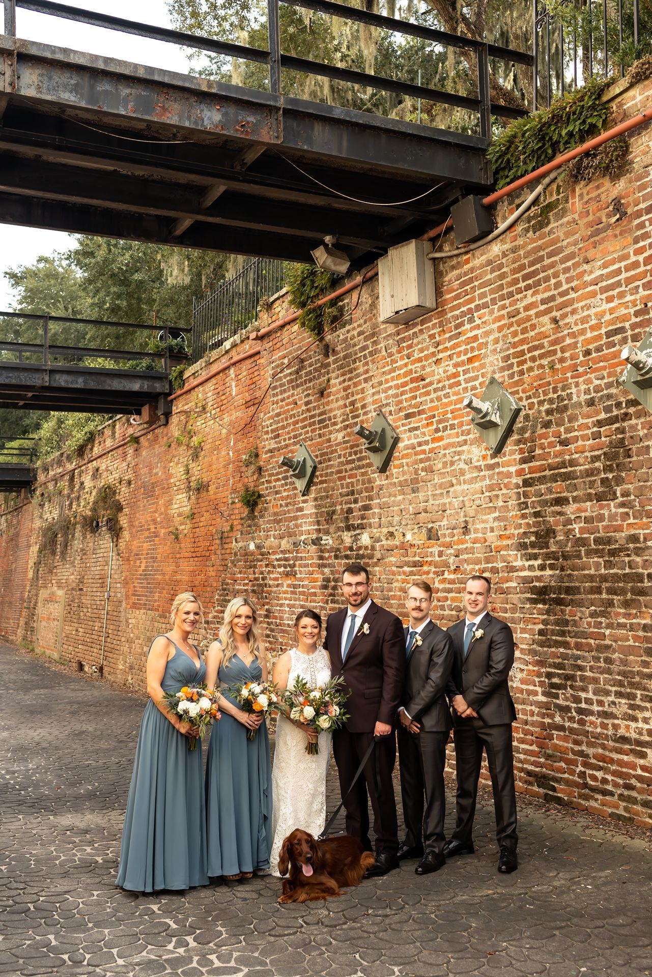 Wedding party poses in front of historic brick wall for formal photos while golden sunlight creates dramatic shadows.