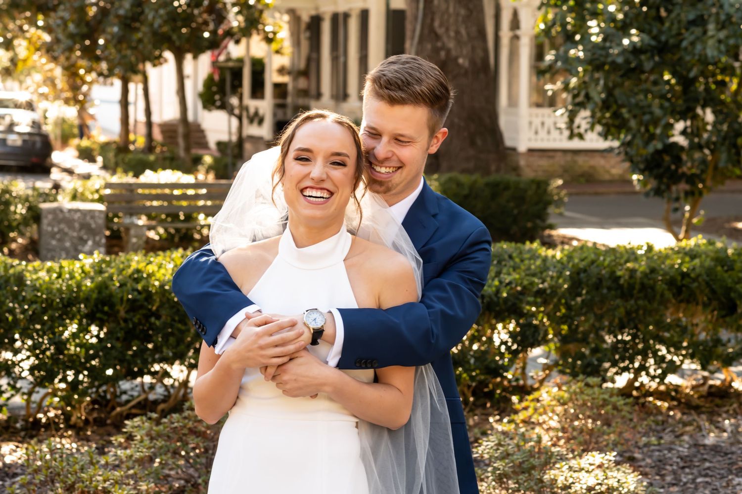 A couple shares a joyful embrace in a garden setting with sunlit trees and greenery in the background.