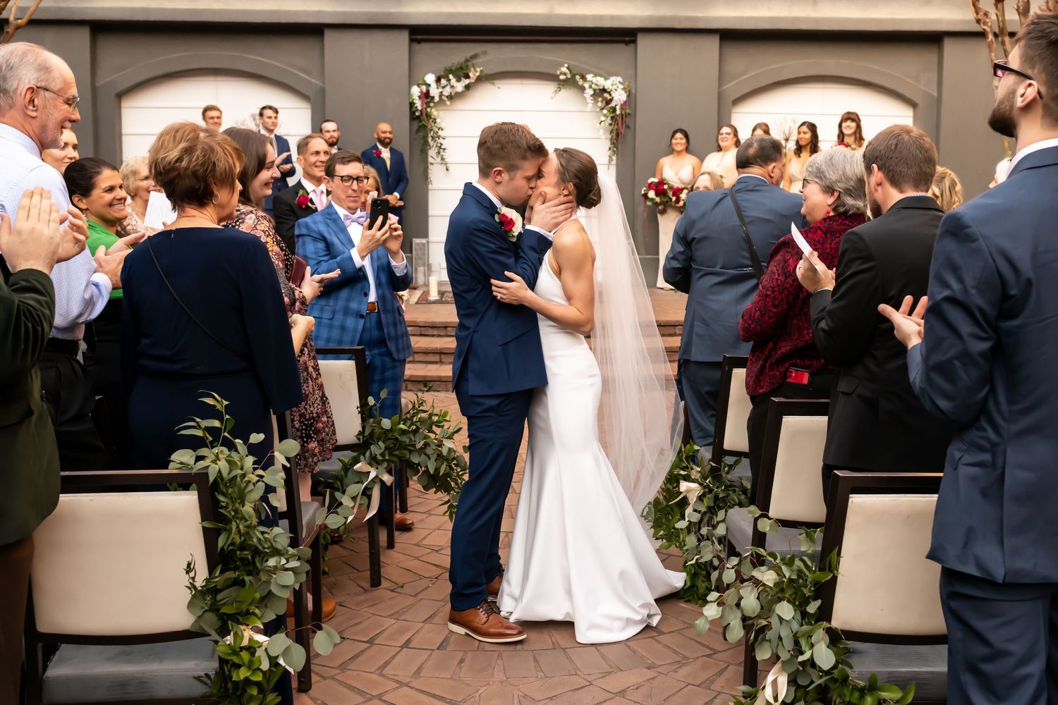 Guests gathered at an elegant wedding reception dance and socialize in a warmly lit indoor venue with decorative greenery.