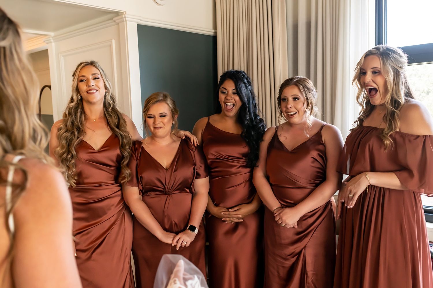 A group of bridesmaids wearing coordinated brown satin dresses pose together in a sunlit room before a wedding.