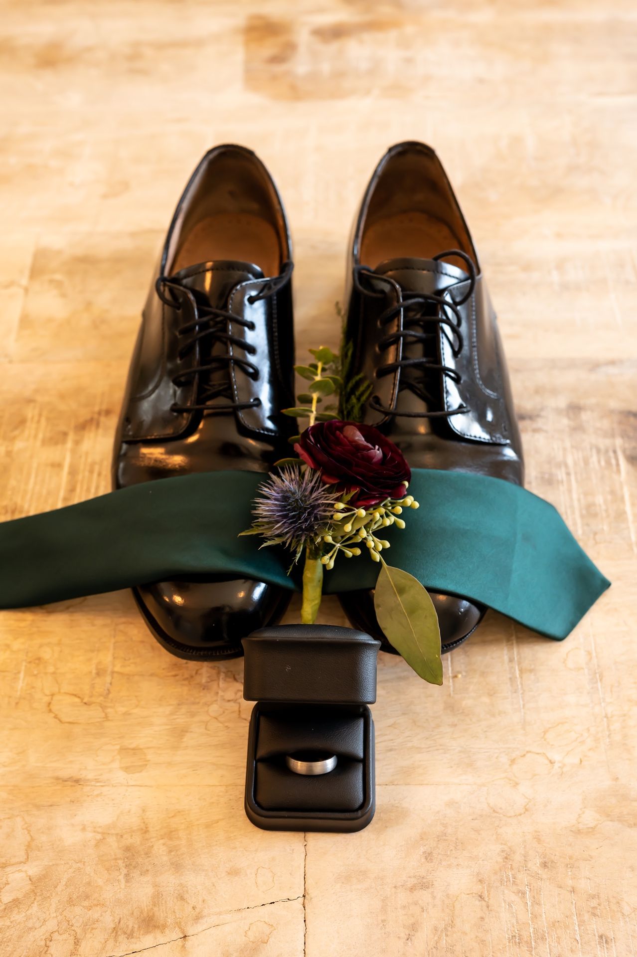 Black patent leather dress shoes with dark green tie and small burgundy floral boutonniere on wooden surface.