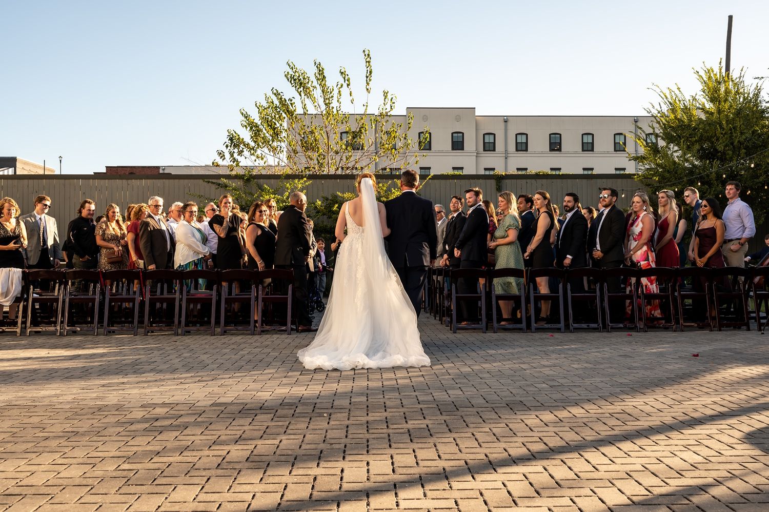 An outdoor wedding ceremony with a large cross and people seated in rows on brick pavement at sunset.