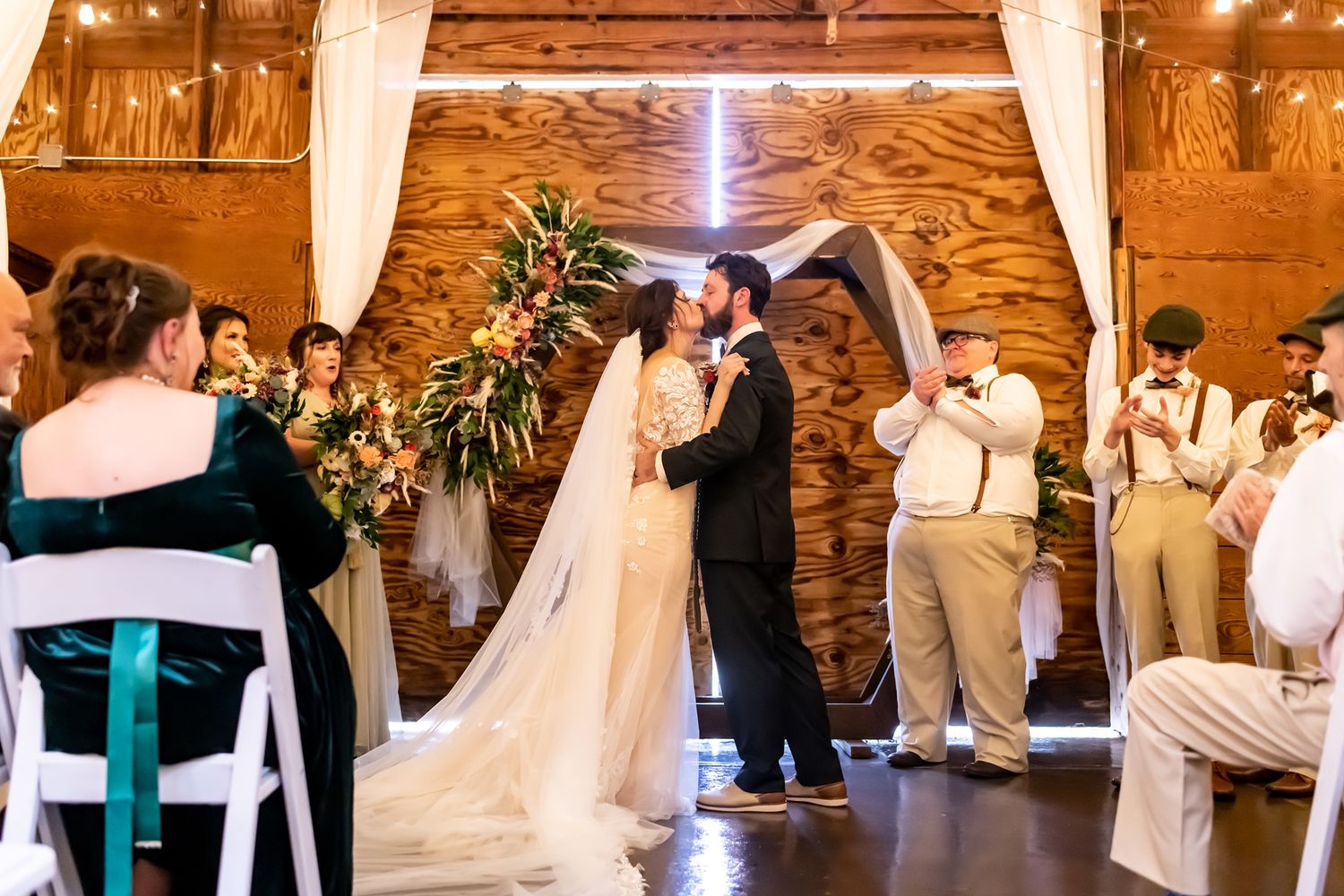A couple shares their first kiss during a rustic barn wedding ceremony with elegant decor and wedding party in attendance.