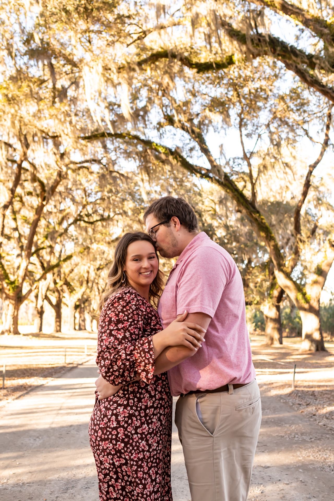 Couple embrace under moss-draped oak trees along a sunlit path in a romantic outdoor setting.