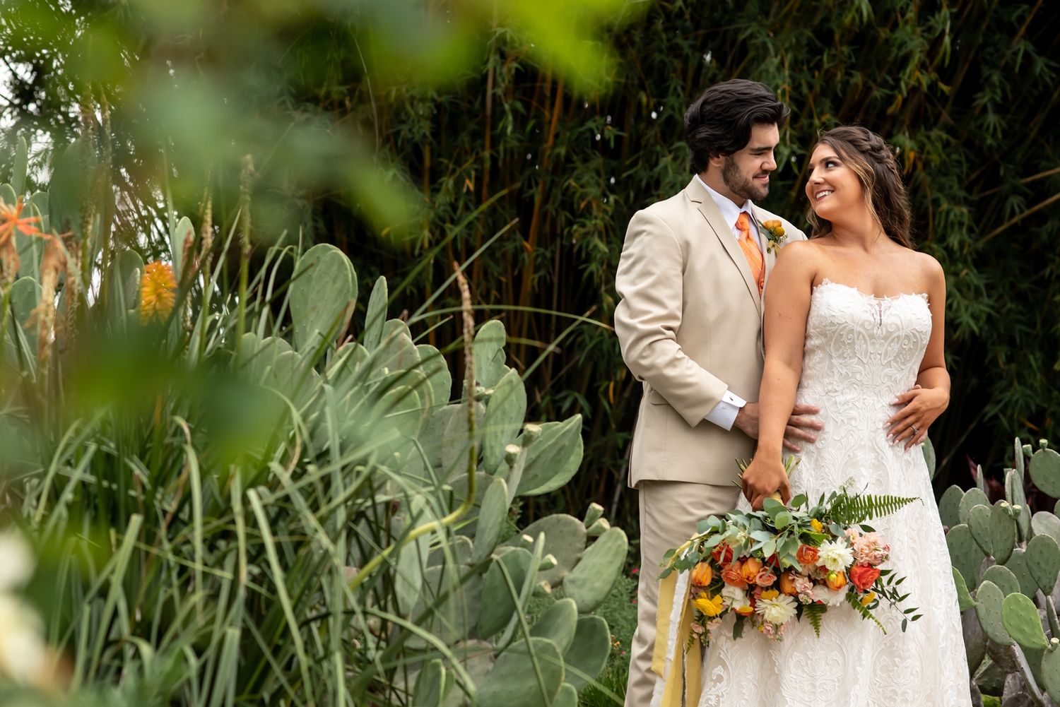 A couple in wedding attire poses together in a lush garden setting with greenery and orange flowers.