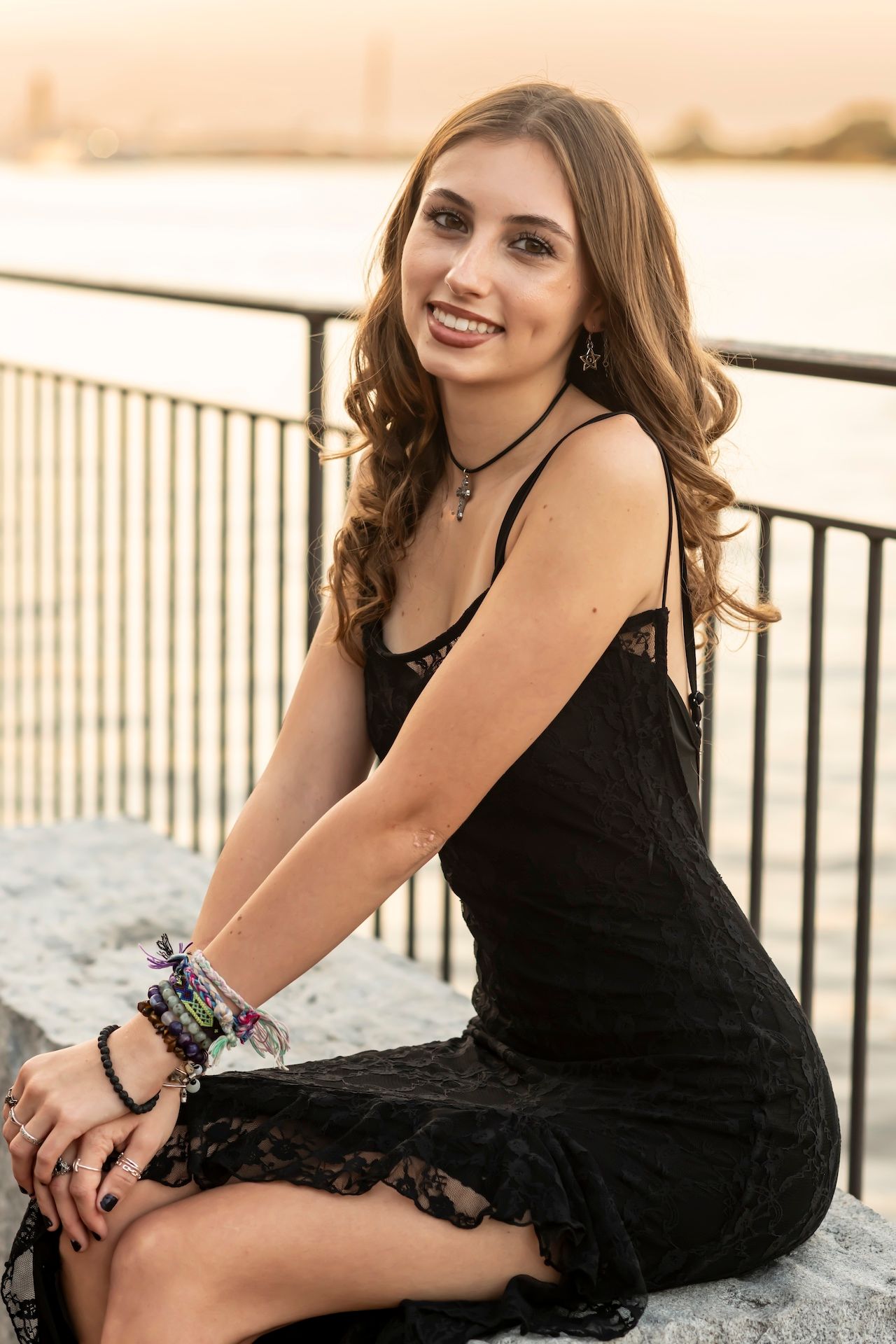 Portrait of someone in a black lace dress sitting on a railing at sunset wearing bracelets and a choker necklace.