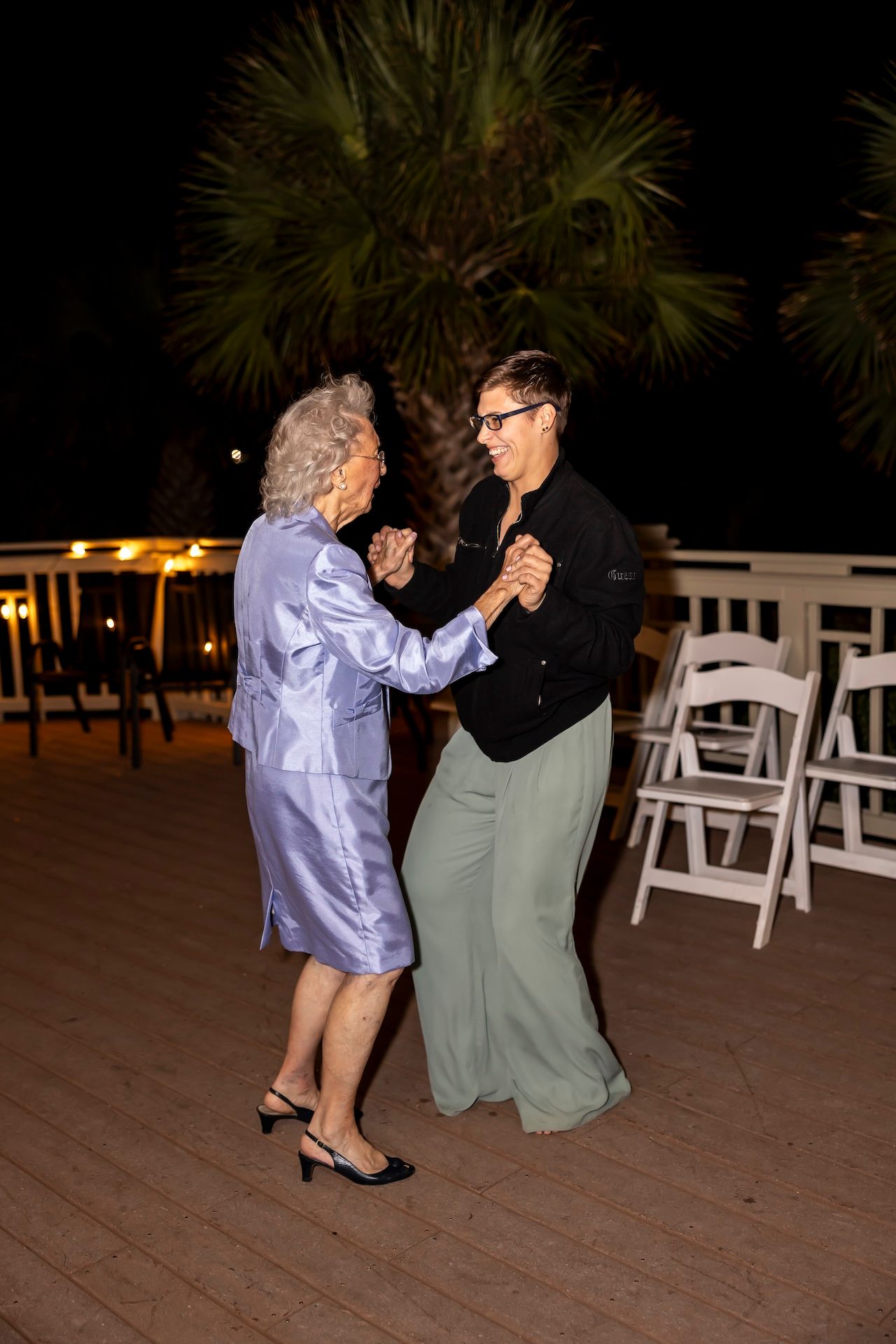Two people dance together at a nighttime event on a wooden deck with white chairs and palm trees in the background.