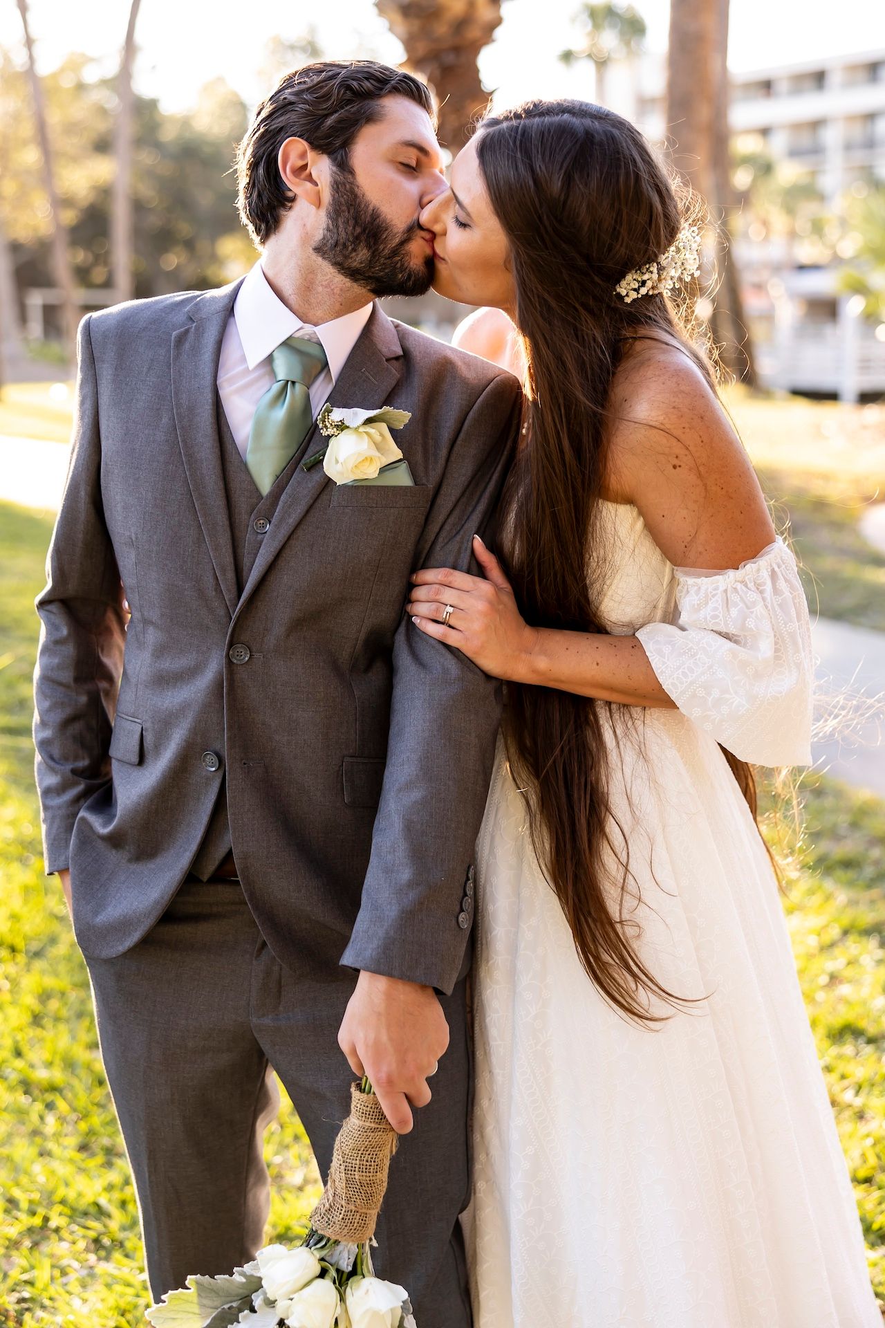 A couple in wedding attire share an intimate kiss outdoors, with the groom in a grey suit and bride in a flowing white dress.
