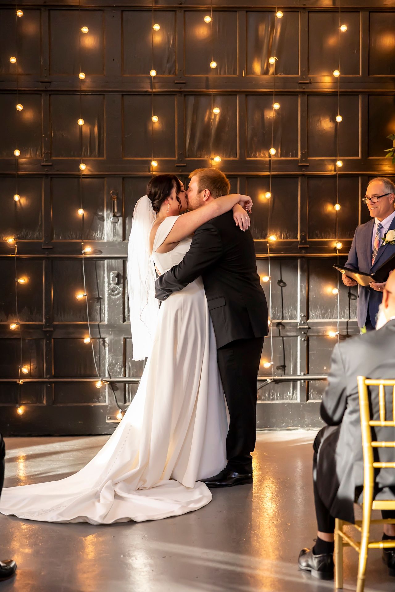 A couple shares their first kiss at the altar with romantic string lights illuminating the industrial-style backdrop.