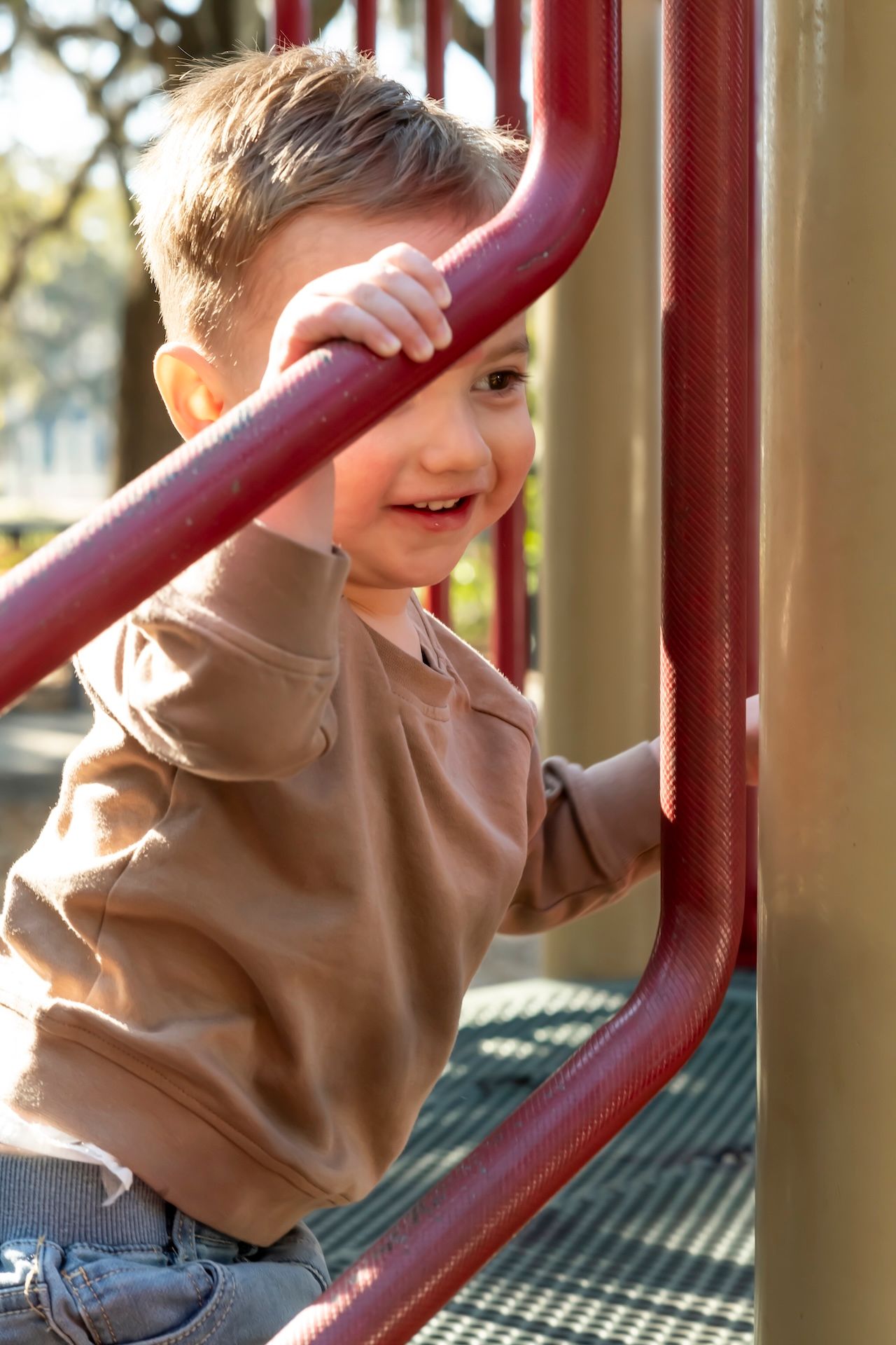 A joyful child plays on playground equipment with burgundy red bars in natural lighting outdoors.