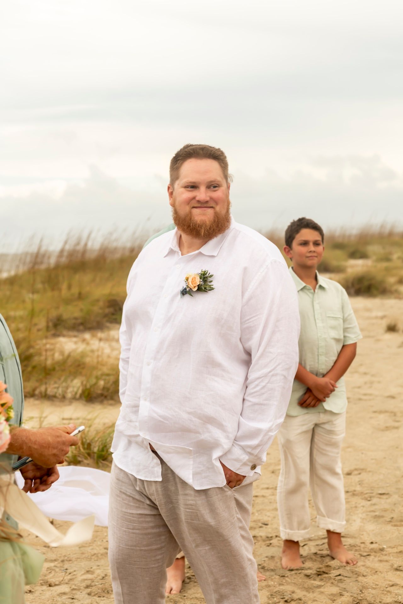 A person wearing a white button-down shirt with a boutonniere stands on a sandy beach while another person waits in background.