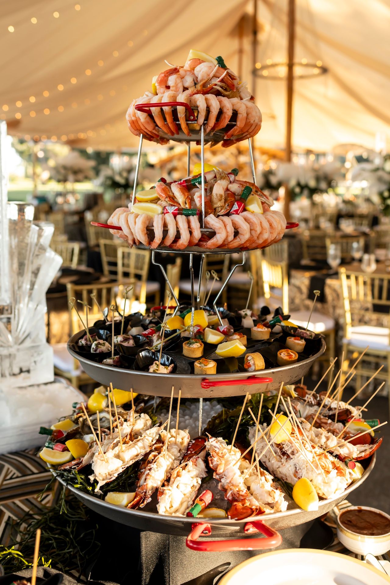 Elegant tiered seafood tower with shrimp cocktail, crab legs and appetizers under a wedding reception tent with string lights.