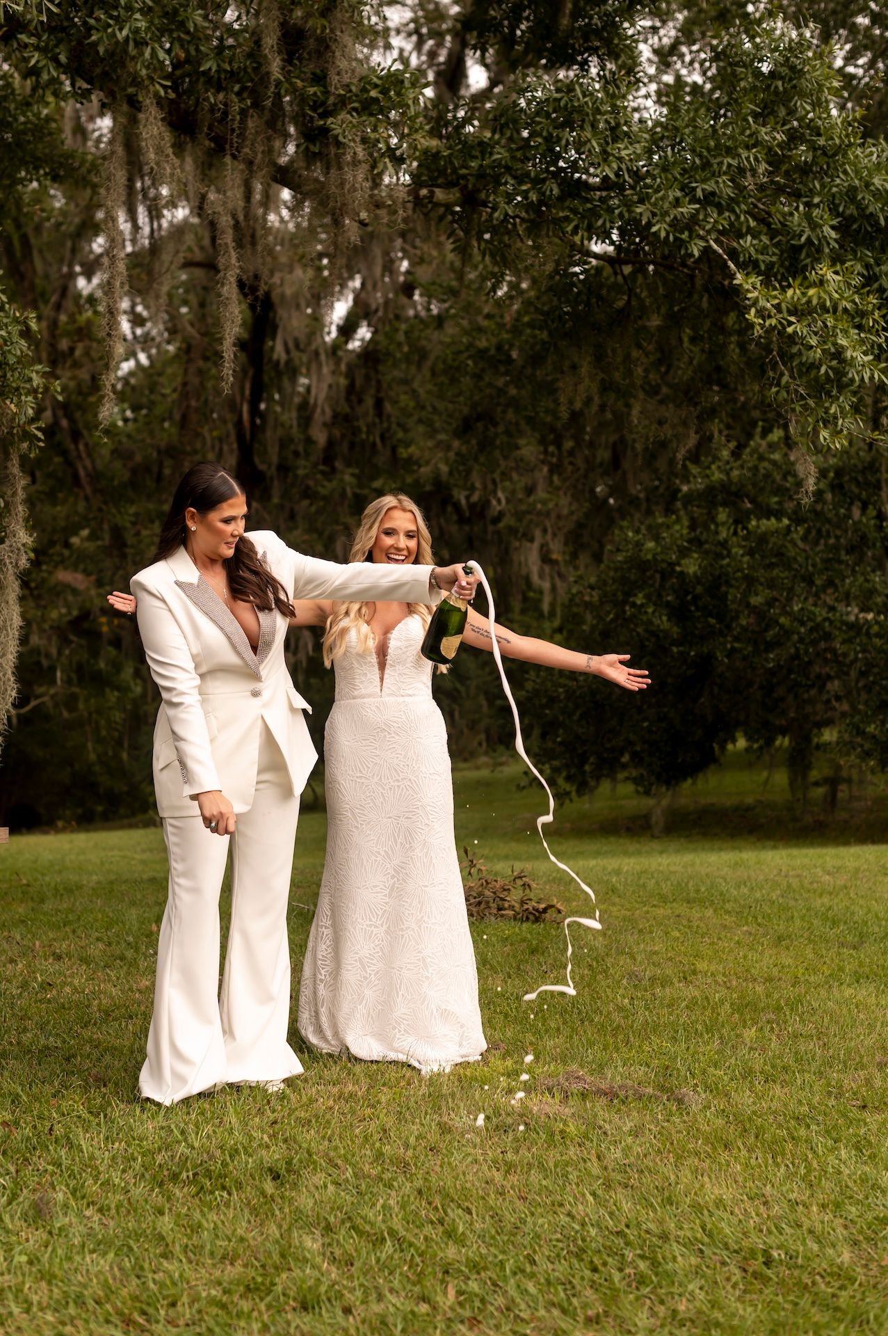 Two people in elegant white outfits celebrate joyfully on a grassy lawn surrounded by Spanish moss-draped trees.