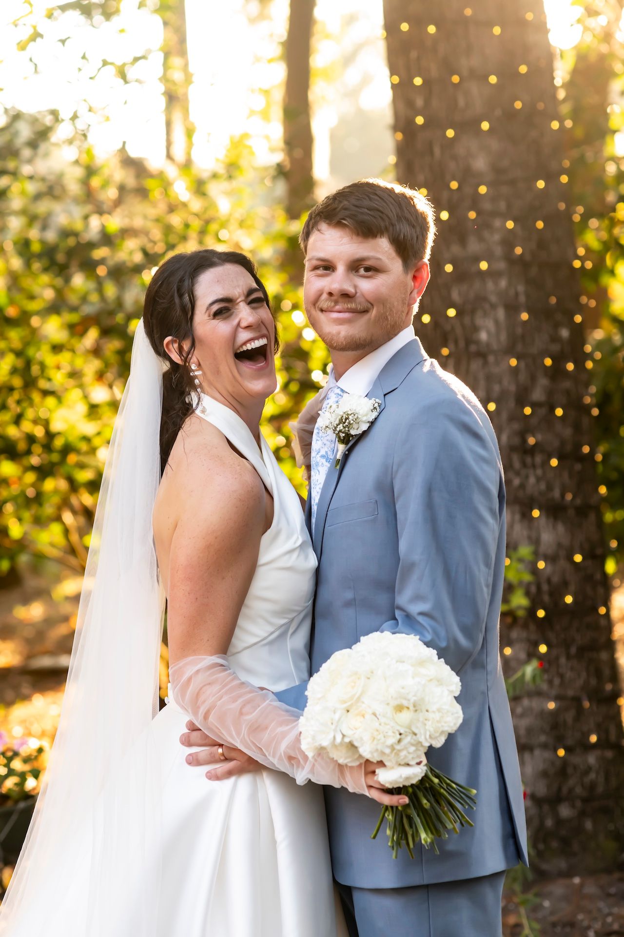 A joyful wedding couple shares a candid moment in front of string lights at sunset.