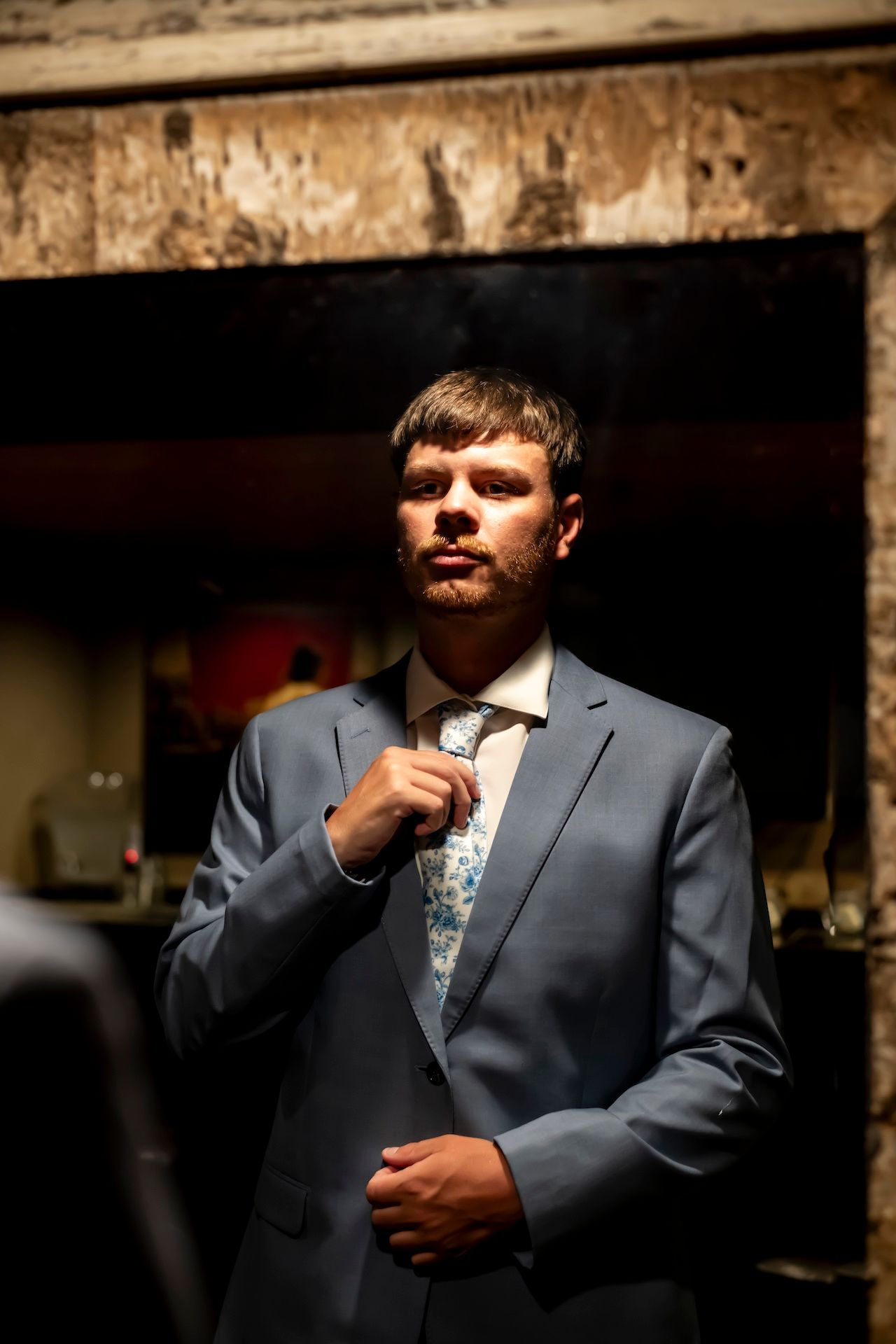 A person adjusts their tie while wearing a gray suit and floral patterned dress shirt in dramatic lighting.