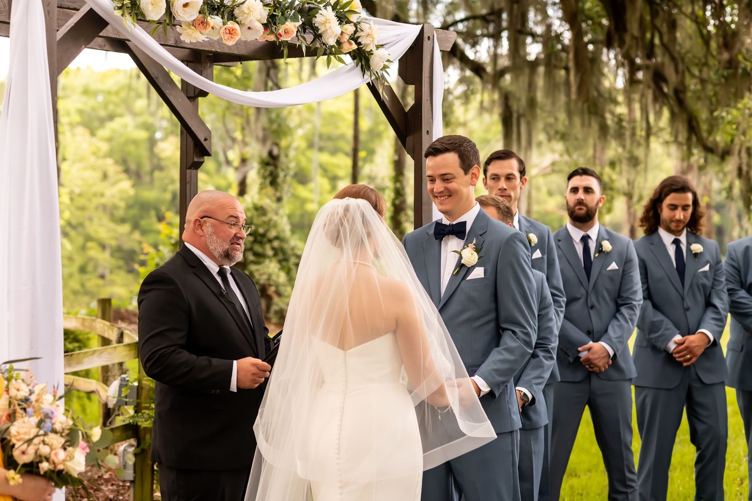 A romantic outdoor wedding ceremony takes place under Spanish moss with groomsmen in gray suits and elegant floral decor.