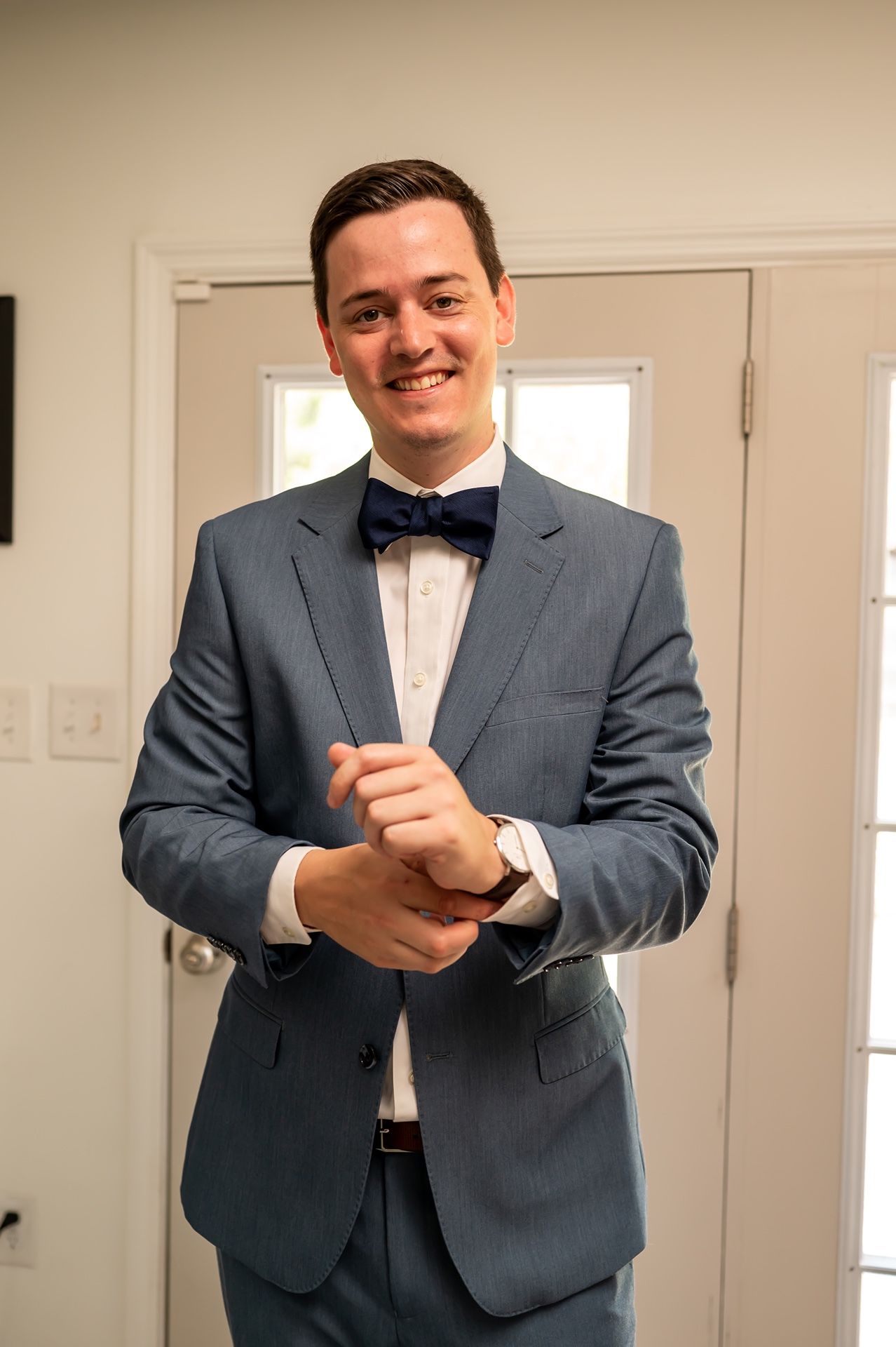 Someone in a gray suit and navy blue bowtie adjusts their cuff while standing in front of a white door.