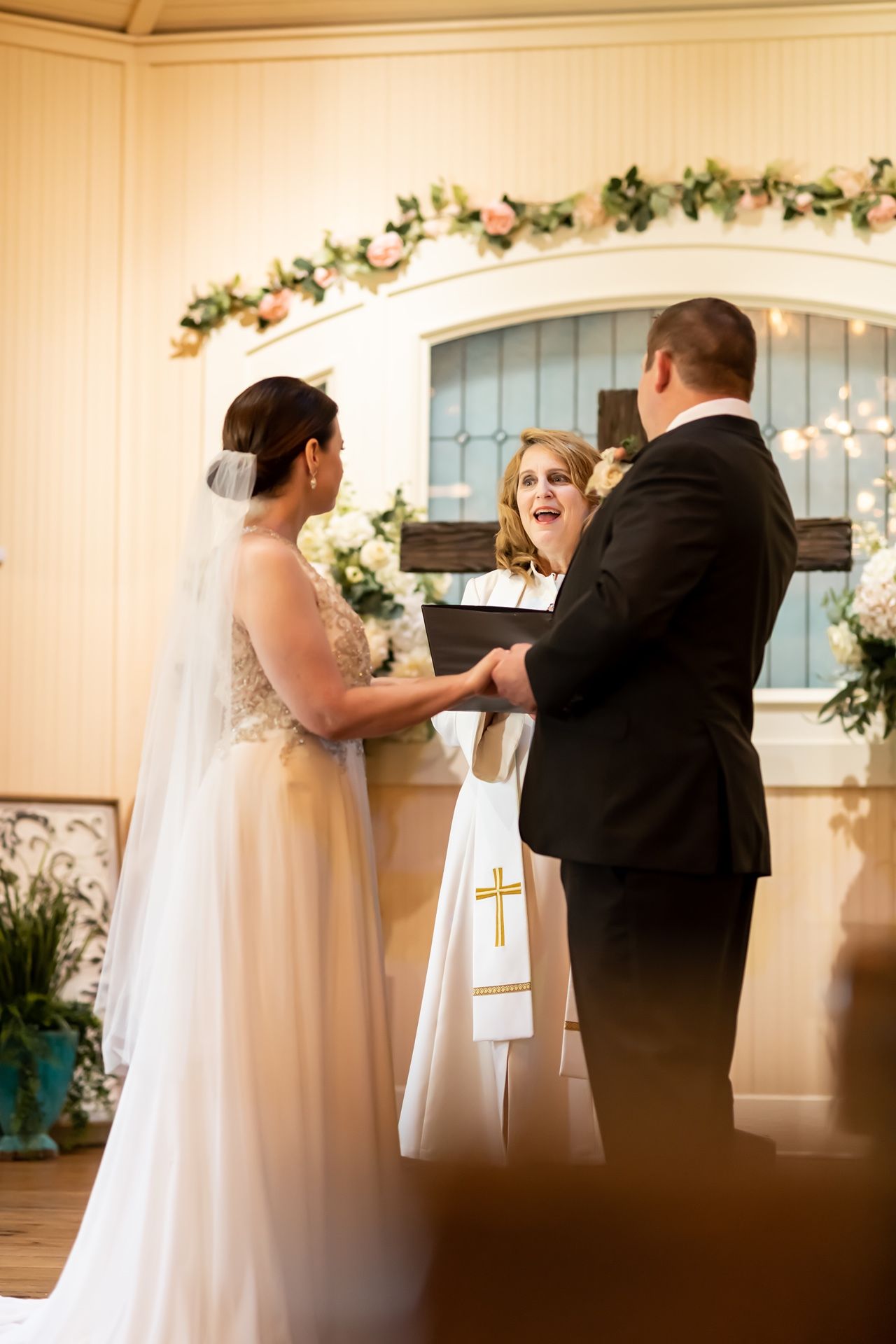 Couple stands at altar with officiant during indoor wedding ceremony decorated with floral garlands and lights.