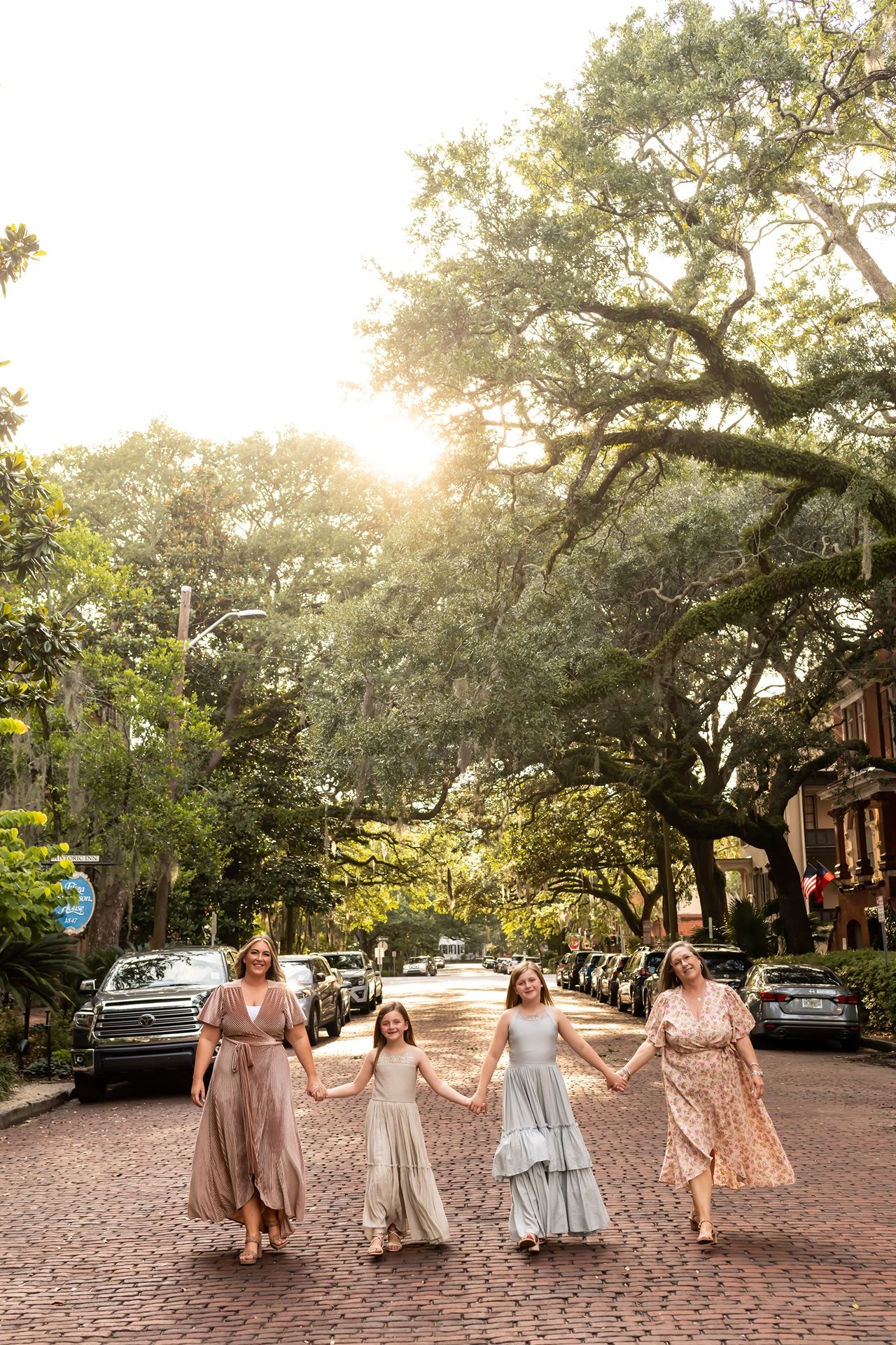 Group in flowy dresses walking together on a brick-paved street under sunlit oak trees in a historic neighborhood.