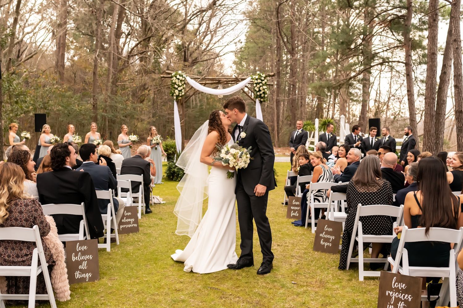 Bride and groom share first kiss at outdoor wedding ceremony with white chairs and rustic wooden arch.