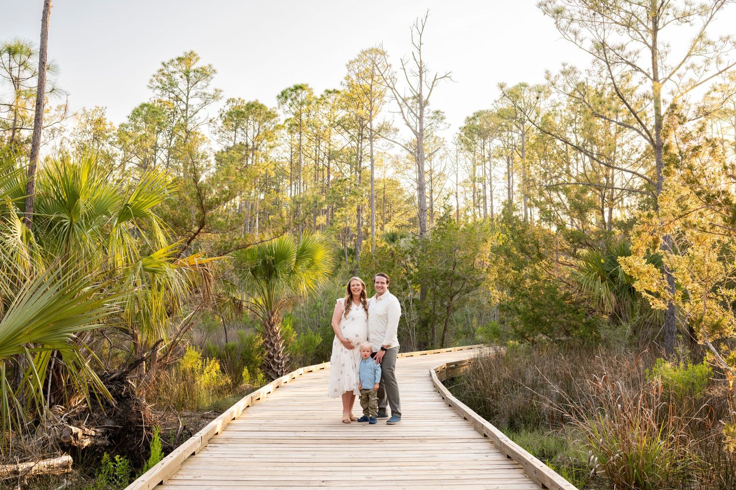 Couple posing on a wooden boardwalk surrounded by lush green trees and palms in a nature preserve.