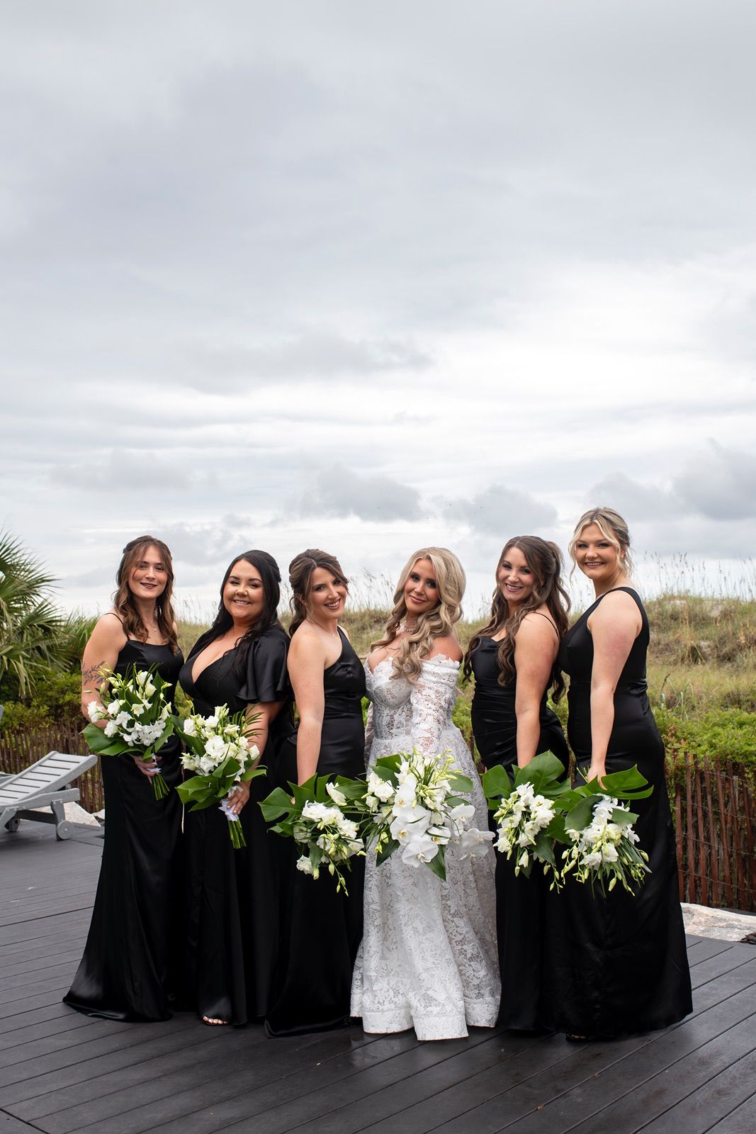 A bridal party in black dresses and white wedding gown pose together on a deck holding white floral bouquets.