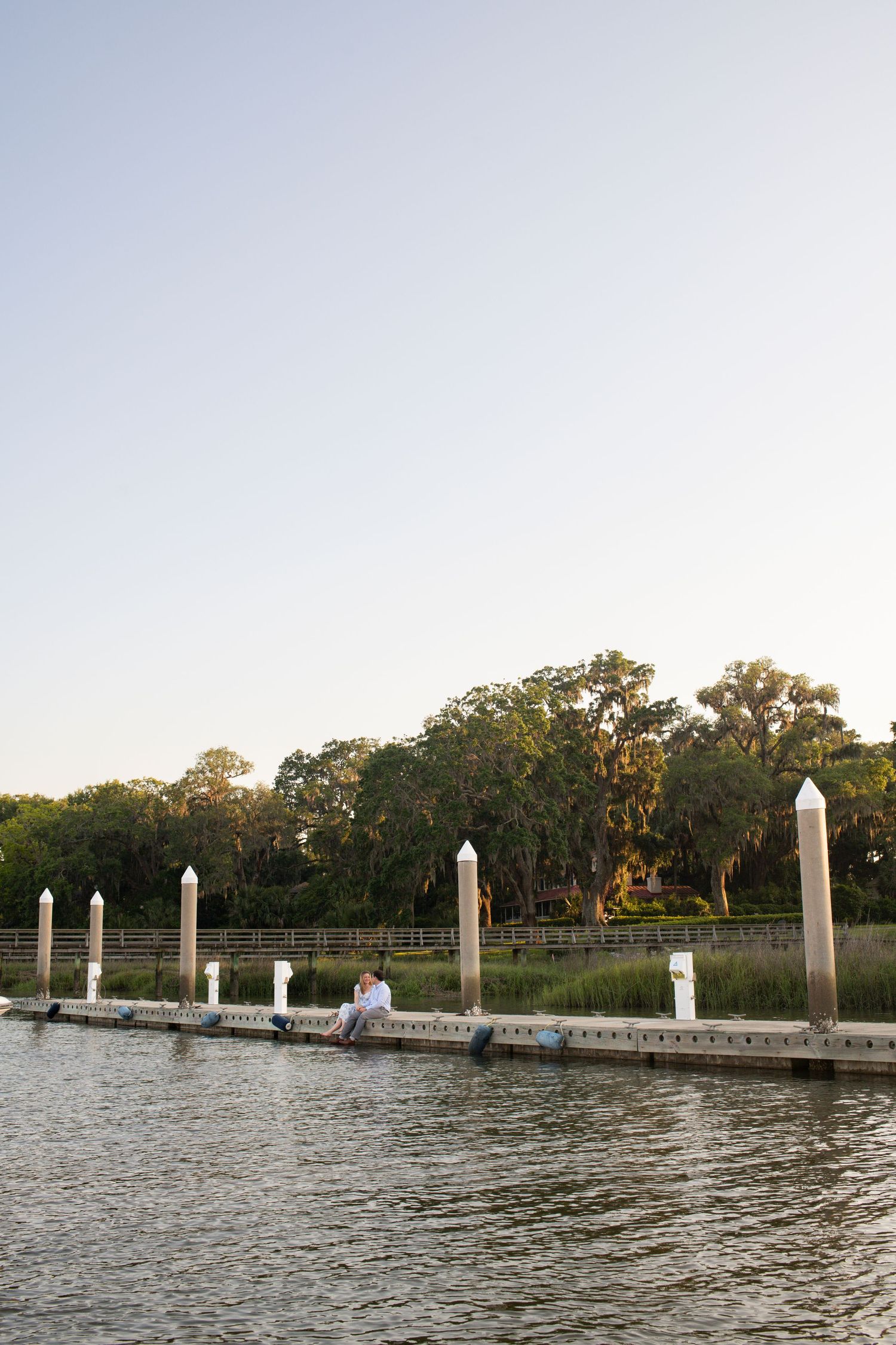 Wooden dock with white posts extends into calm waters at sunset with trees lining the distant shore.