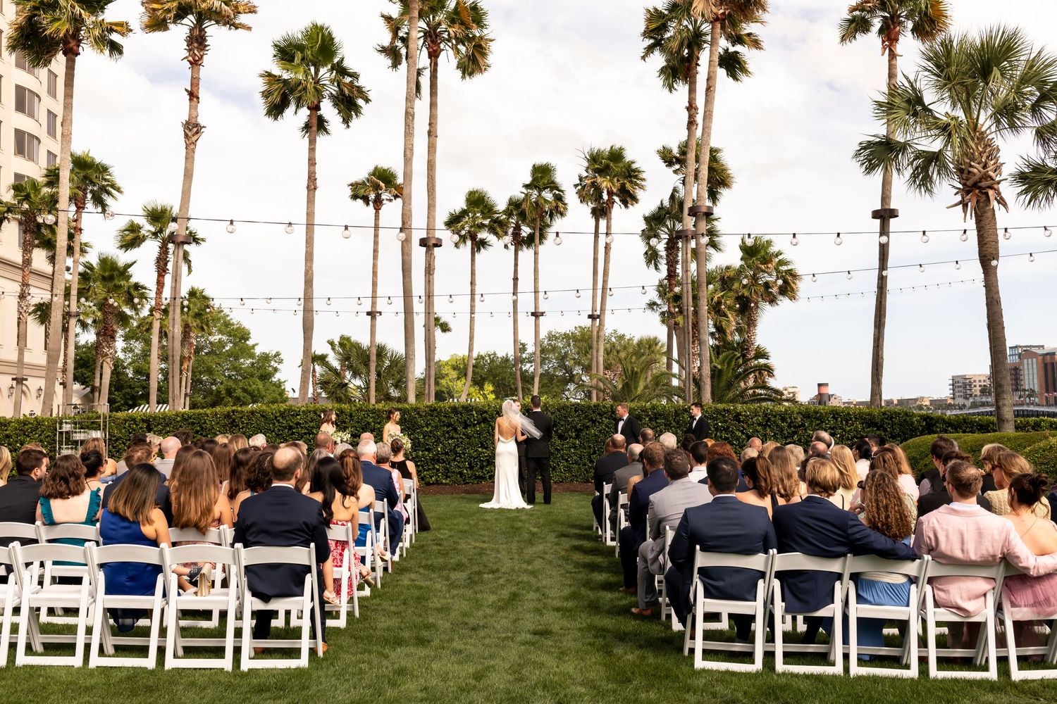 Outdoor wedding ceremony setup with white chairs and tall palm trees against blue sky.