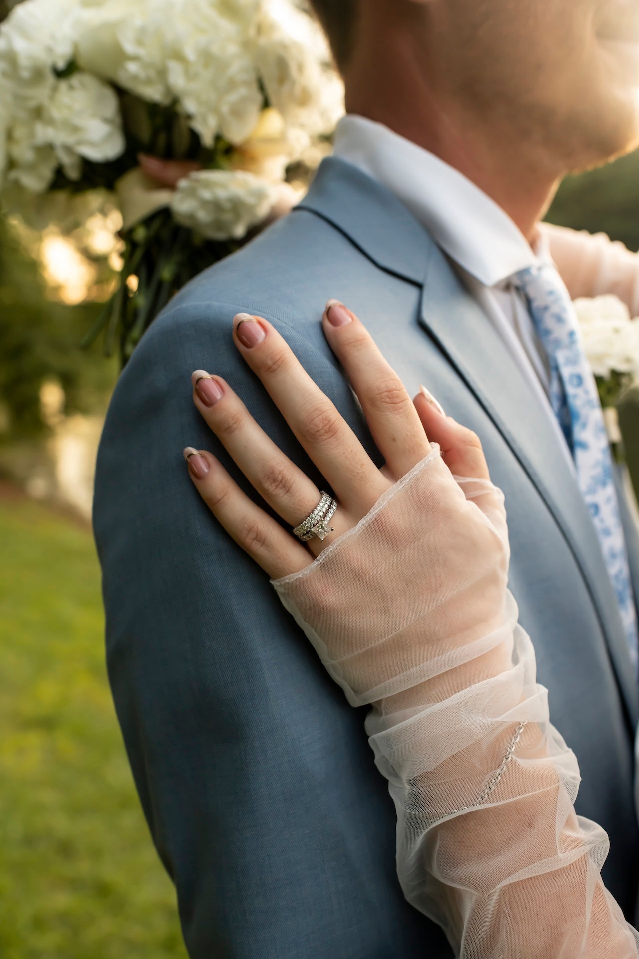 A close-up of an elegant sheer-sleeved hand with wedding rings resting on a light blue suit jacket.