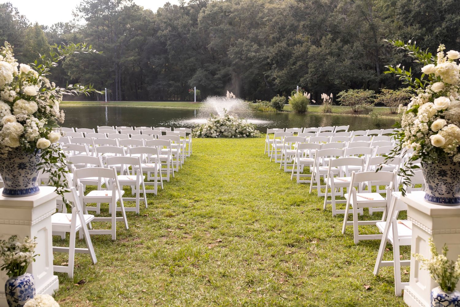 White wedding ceremony setup with rows of chairs and floral arrangements leading to a fountain in a garden setting.