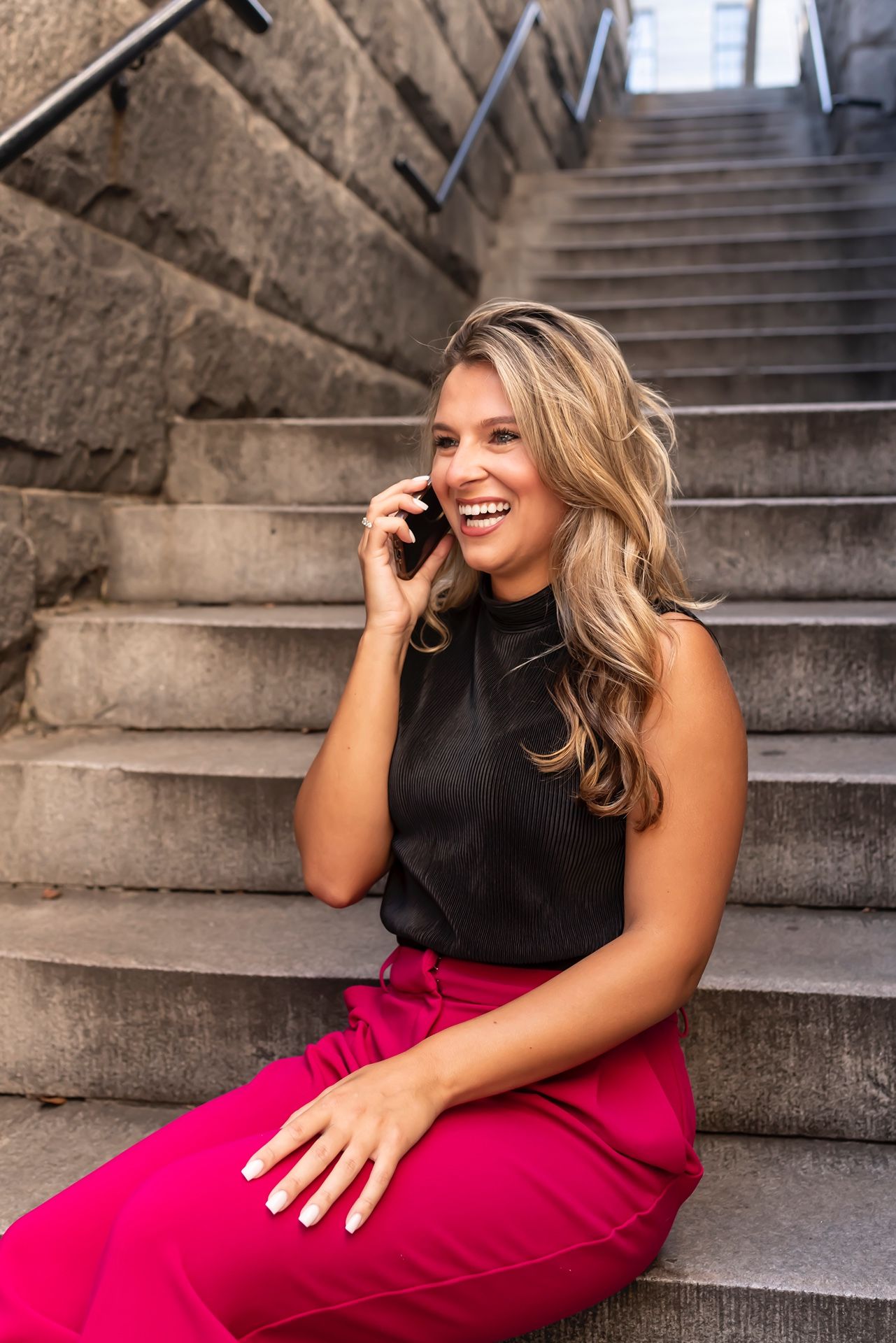 Person in black sleeveless top and vibrant red pants sits on concrete stairs while talking on phone and smiling.