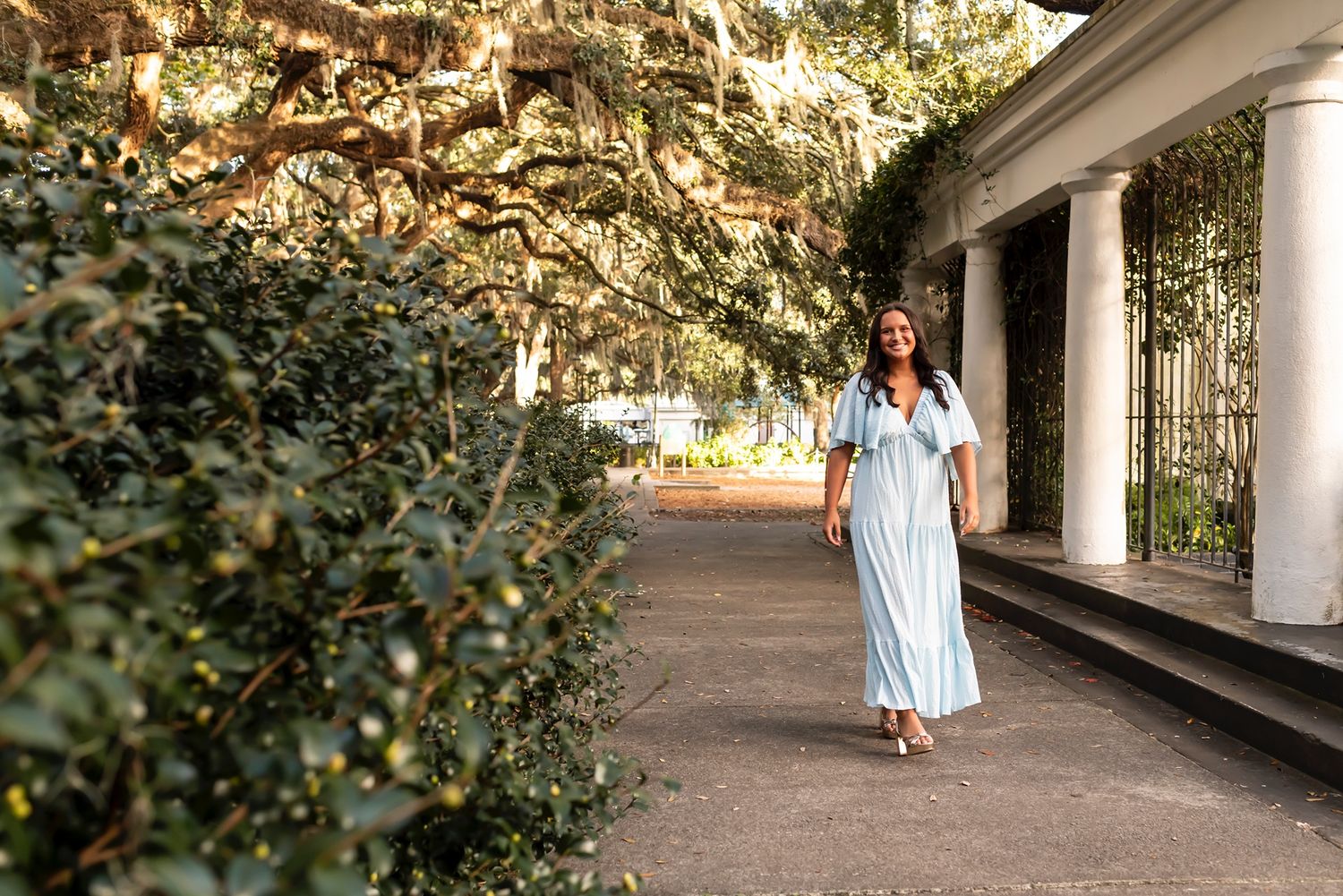 Person in flowing white dress walks along covered outdoor pathway surrounded by lush greenery in warm sunlight.